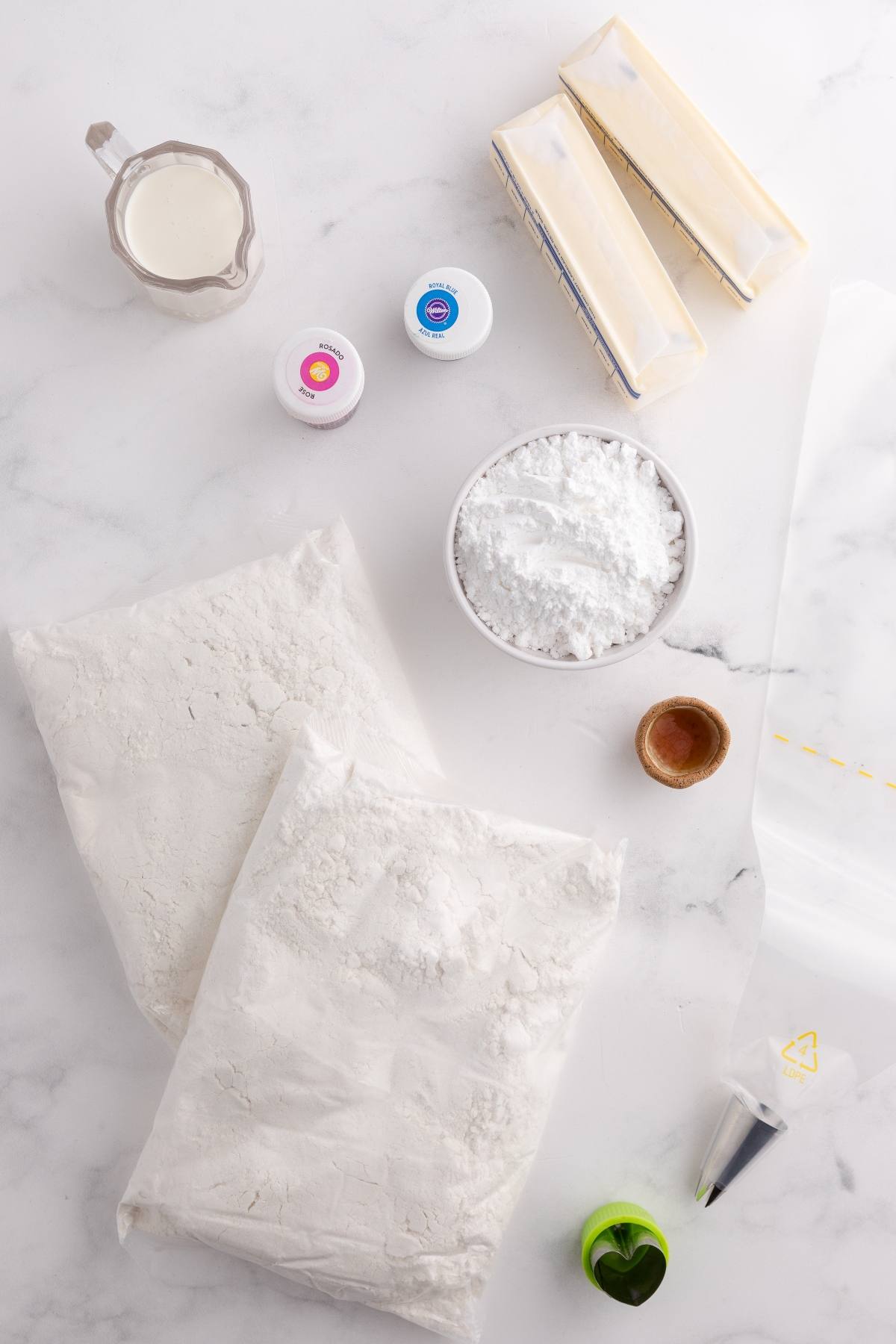 Baking ingredients on a white surface: flour, butter, milk, powdered sugar, food color, piping tip, and a small bowl.