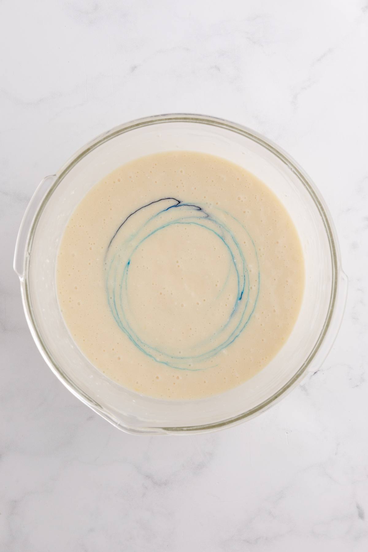 Glass bowl with pancake batter and a swirl of blue food coloring on a white marble surface.