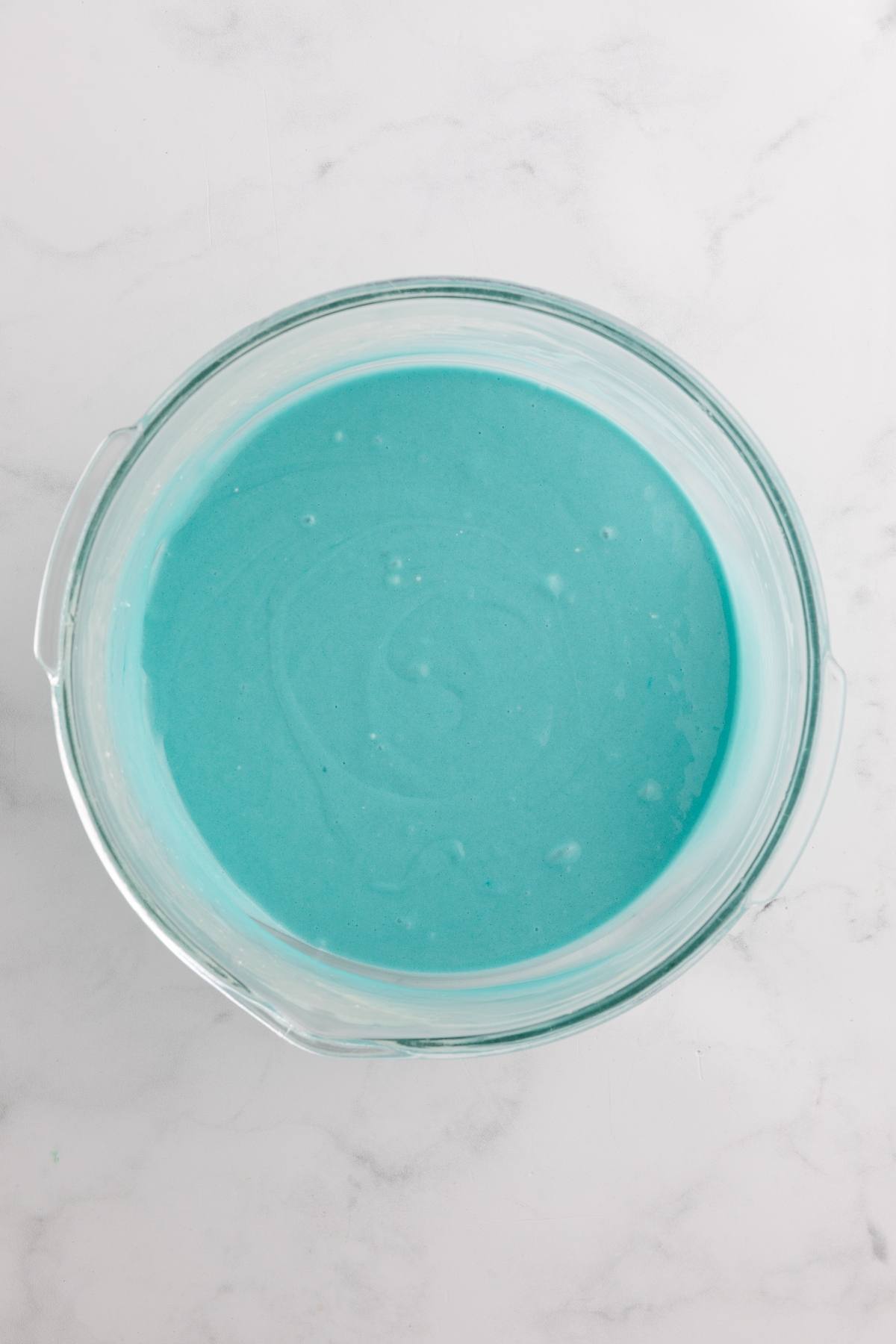 A glass bowl filled with smooth, light blue cake batter on a white marble surface.