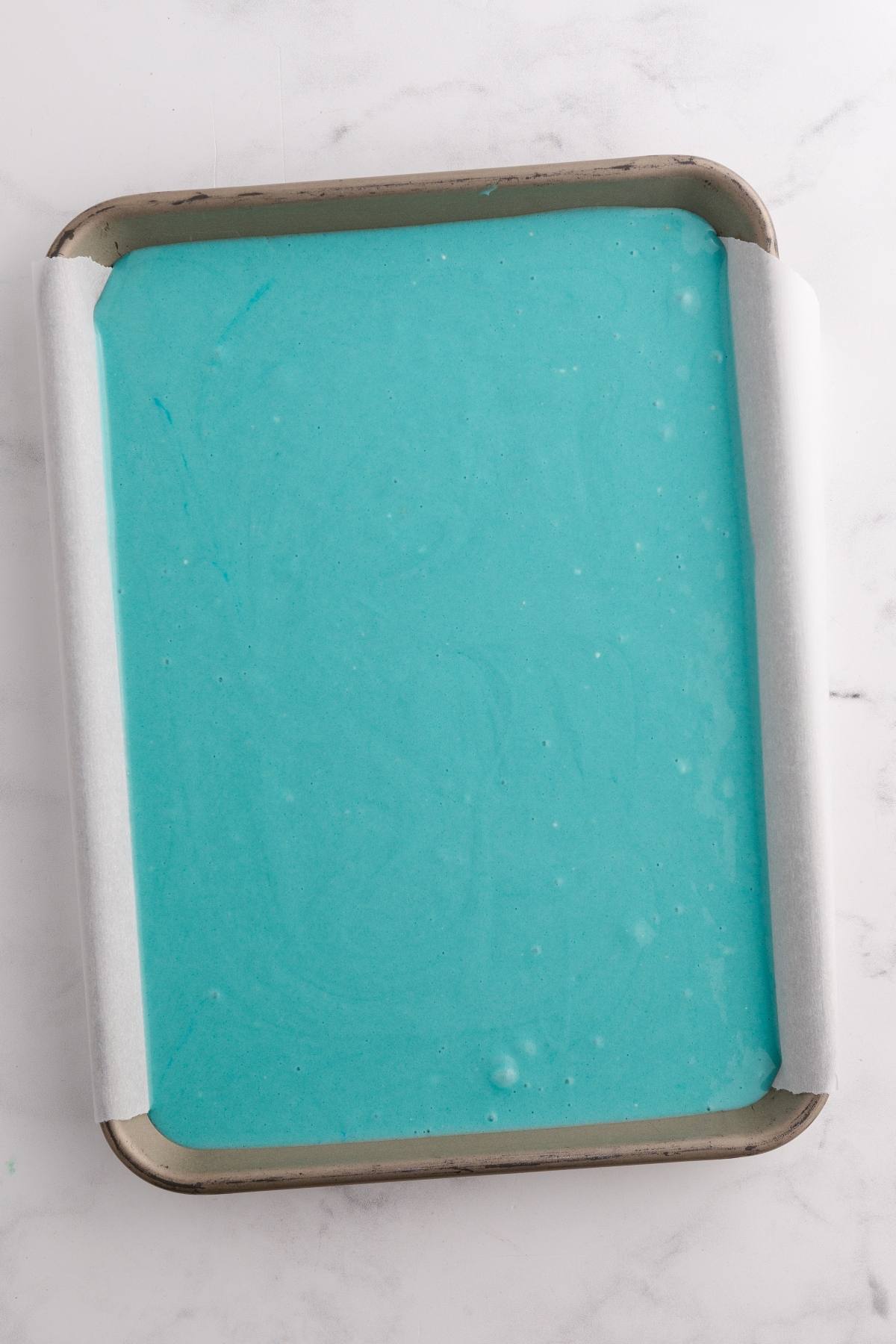 A baking tray lined with parchment paper filled with smooth, bright blue batter on a marble surface.