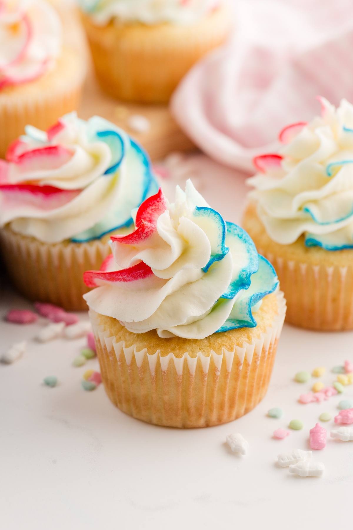 Vanilla gender reveal  cupcakes with swirled red, white, and blue frosting, surrounded by pastel sprinkles.