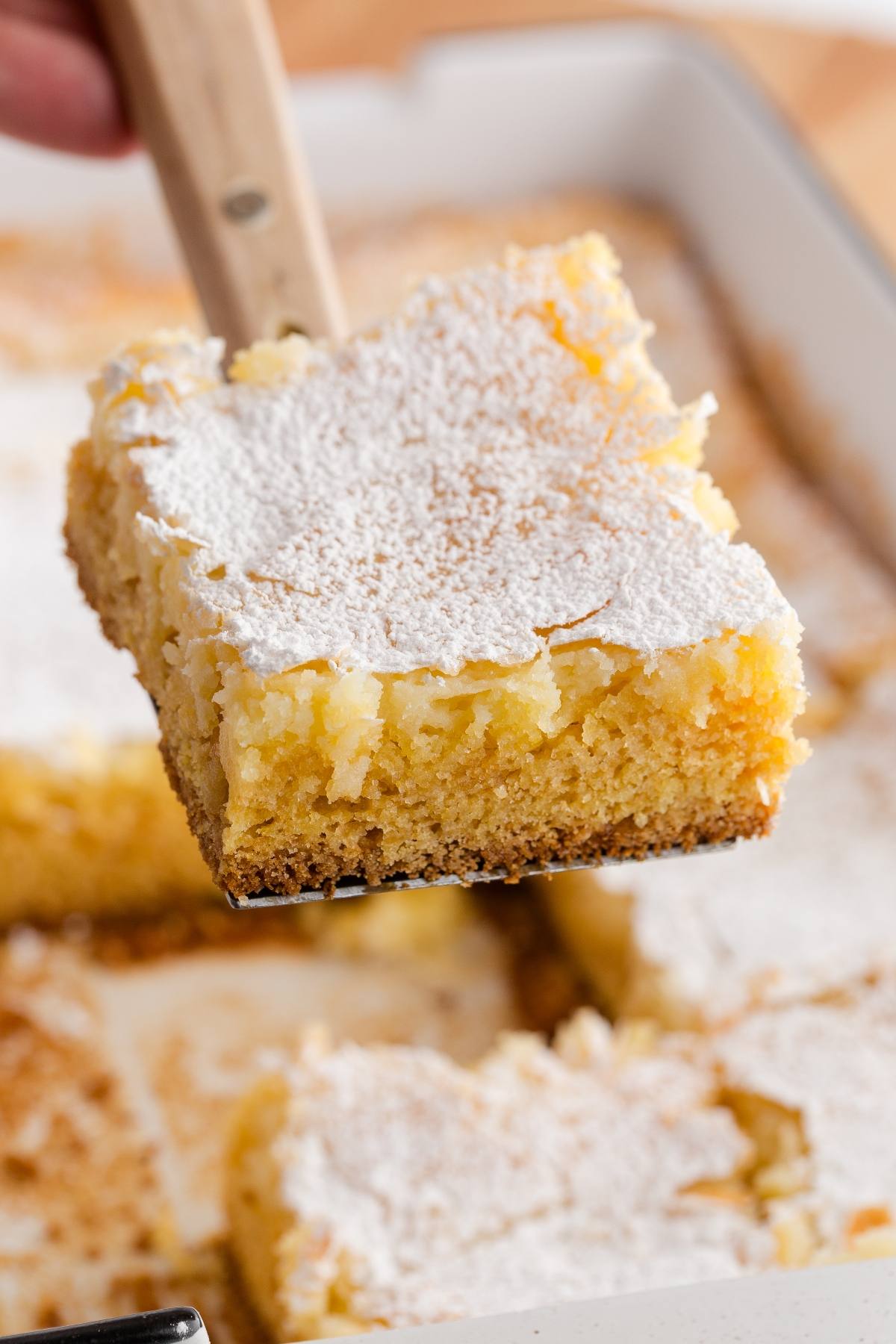 A spatula lifts a square piece of gooey butter cake dusted with powdered sugar from a baking dish.