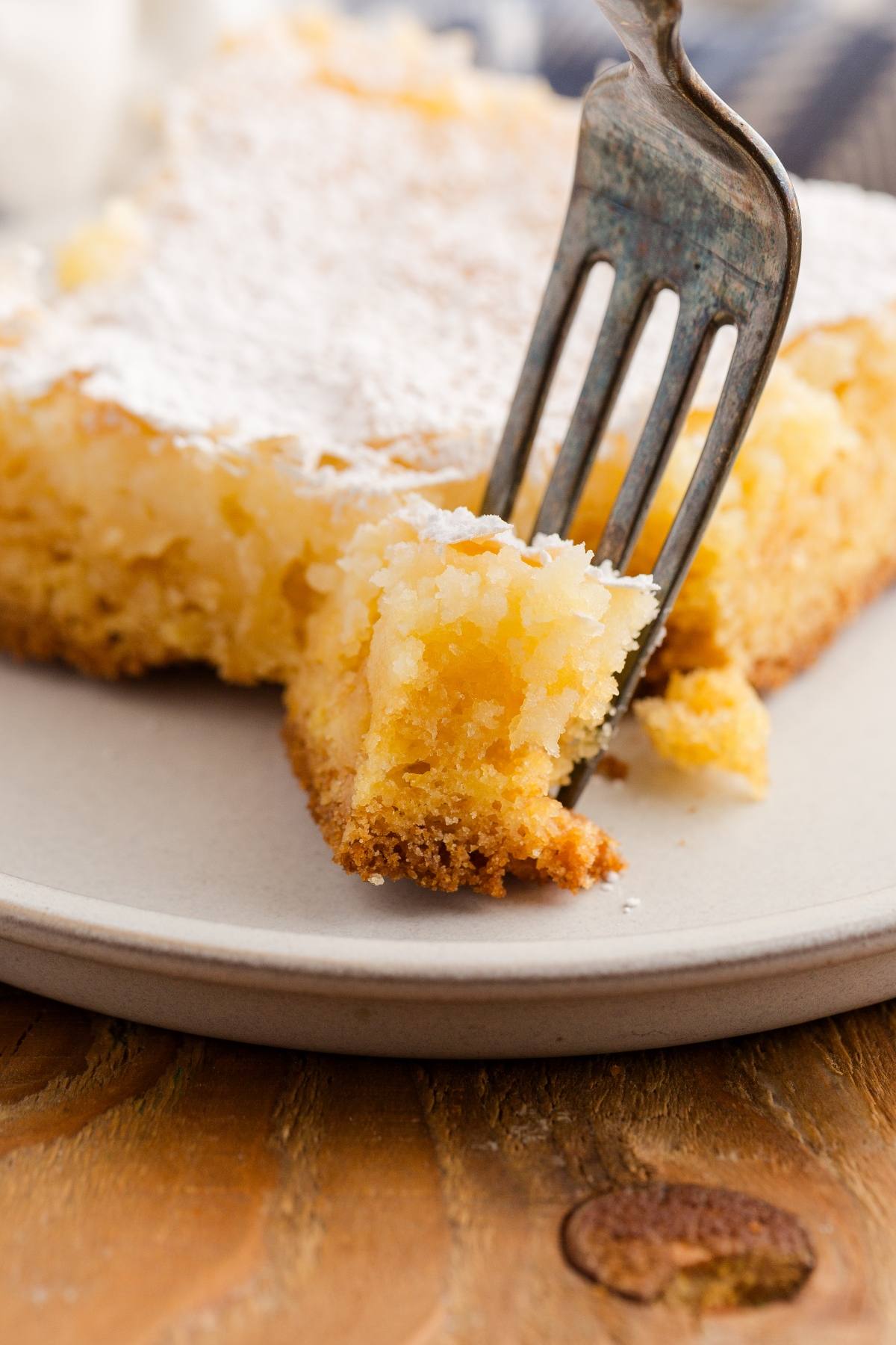 A fork cutting into a powdered sugar-topped gooey butter cake on a white plate.