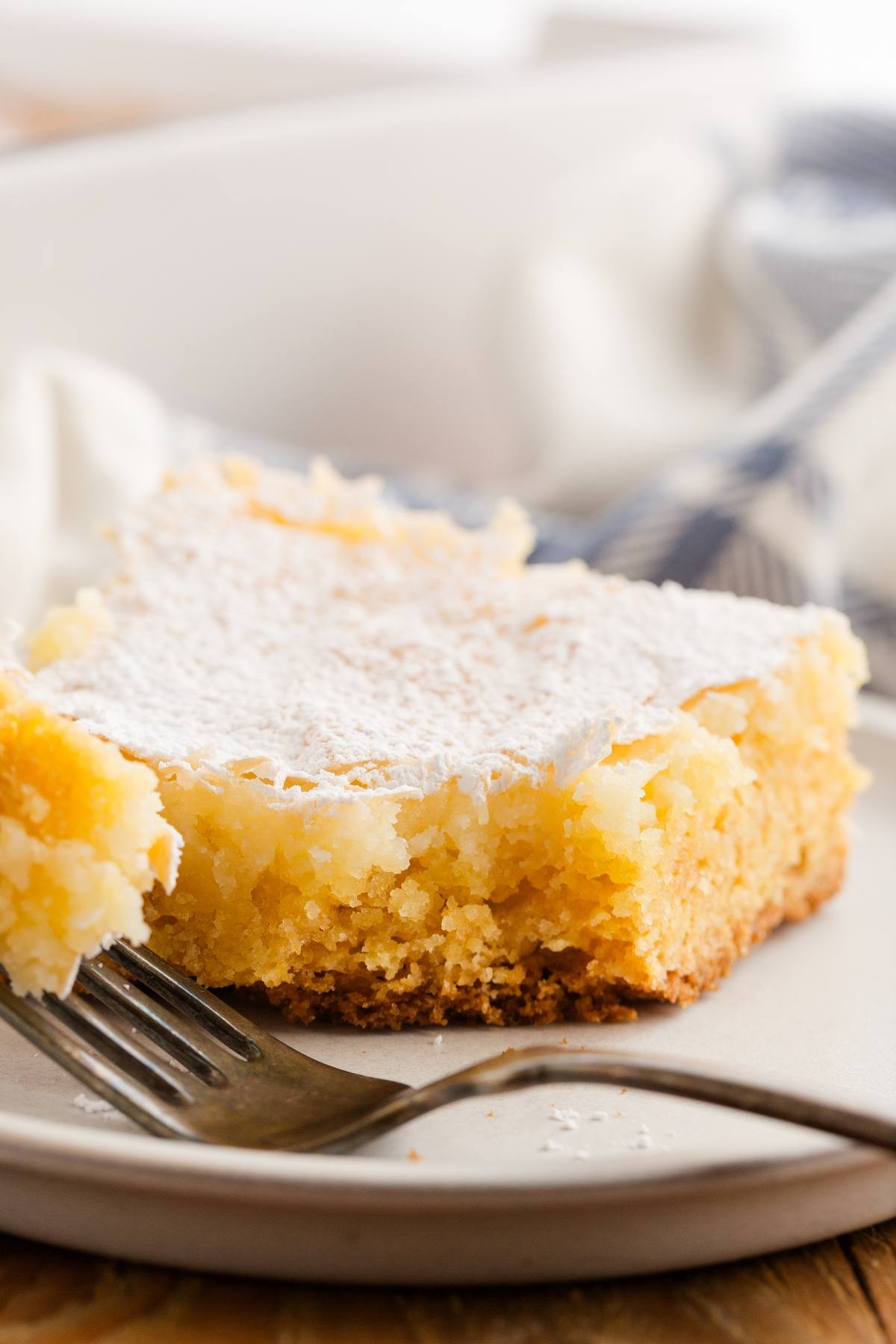 A close-up of a gooey butter cake slice on a plate with a fork, dusted with powdered sugar.