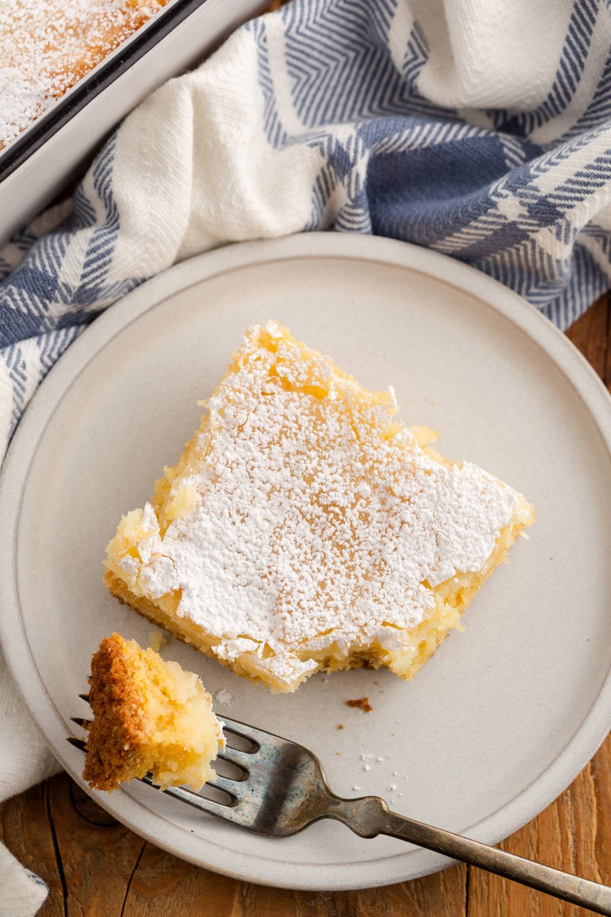 A square slice of powdered sugar-topped cake on a plate, with a fork holding a small bite.