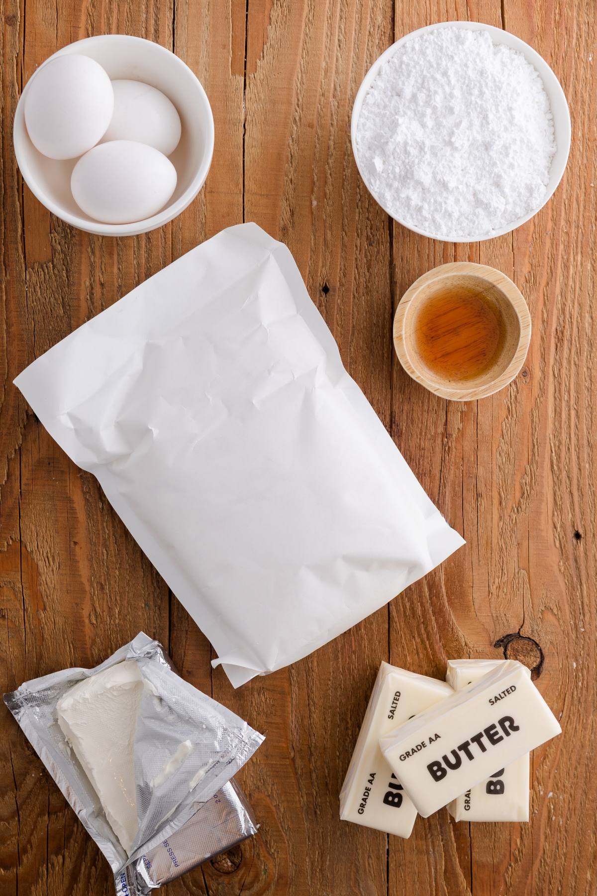 Baking ingredients on a wooden table: eggs, flour, powdered sugar, vanilla, cream cheese, and butter.