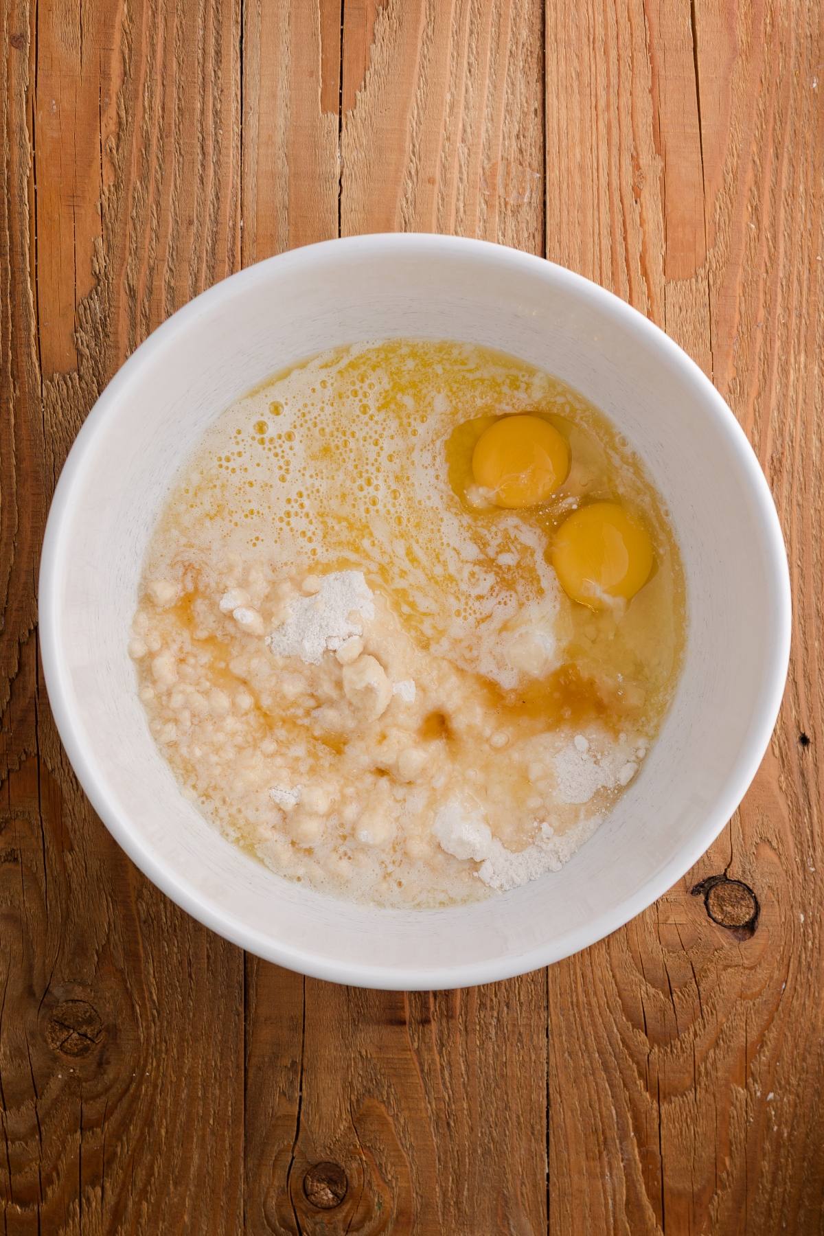 White bowl with eggs, flour, sugar, and liquid ingredients on a wooden surface, ready to be mixed.
