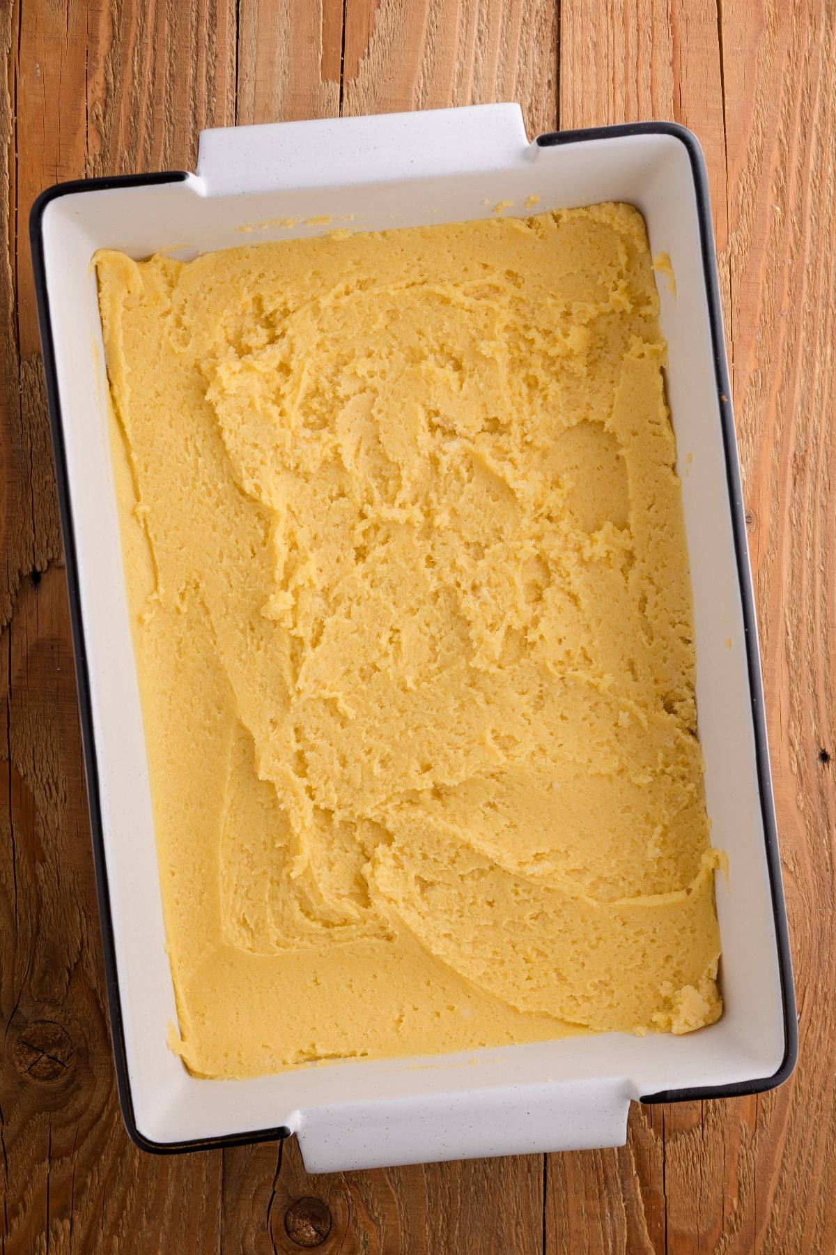 A baking dish filled with smooth, uncooked cornbread batter on a wooden surface.