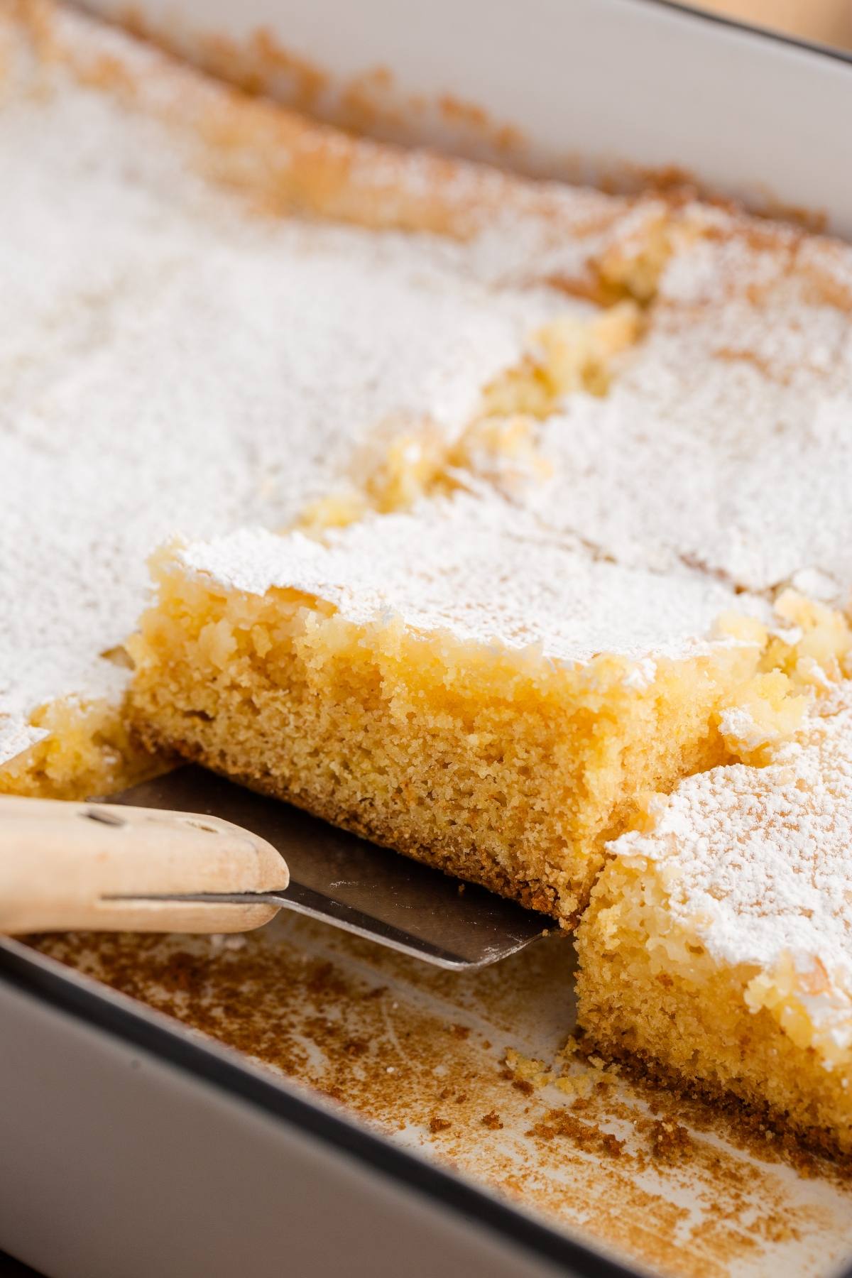 A spatula lifts a powdered sugar-topped lemon bar from a baking dish of sliced lemon bars.