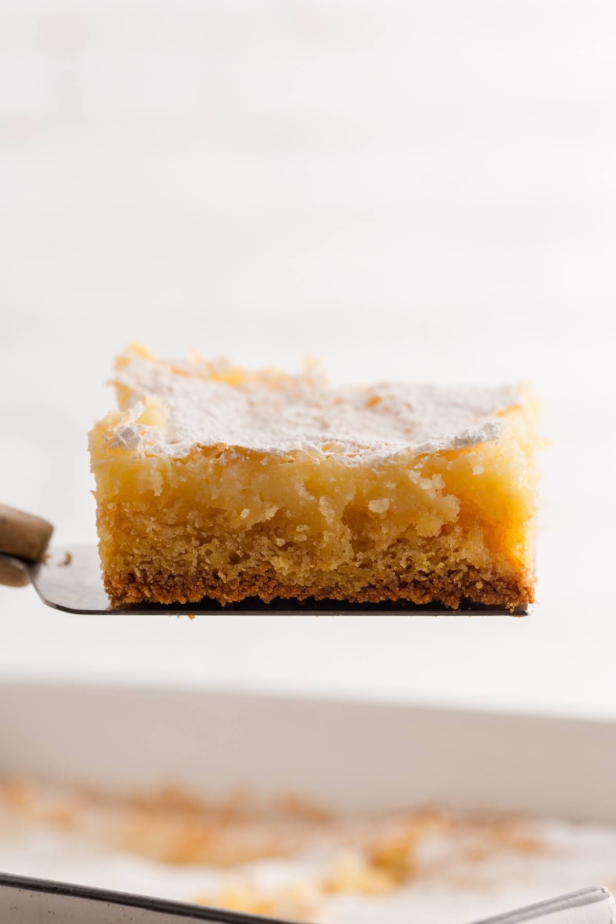 A gooey butter cake square on a spatula, showing a golden, moist, and dense texture with a powdered sugar top.