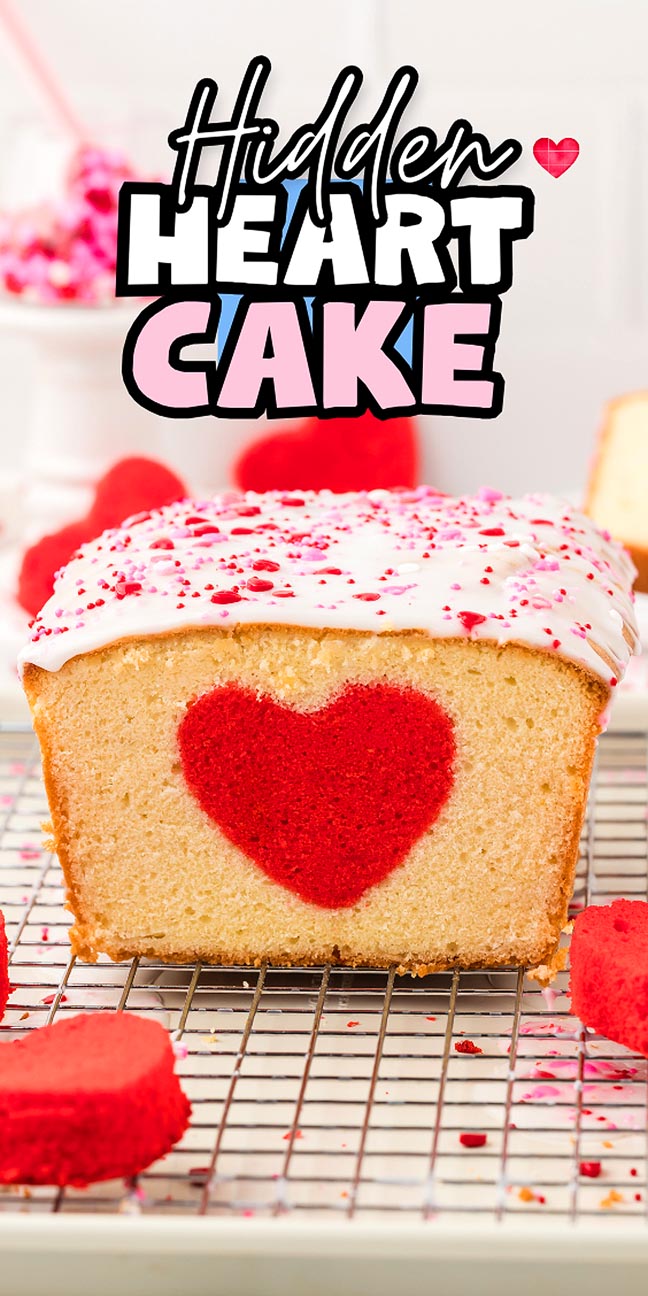 A hidden heart cake with a red heart shape inside, topped with white icing and sprinkles, sits on a cooling rack.