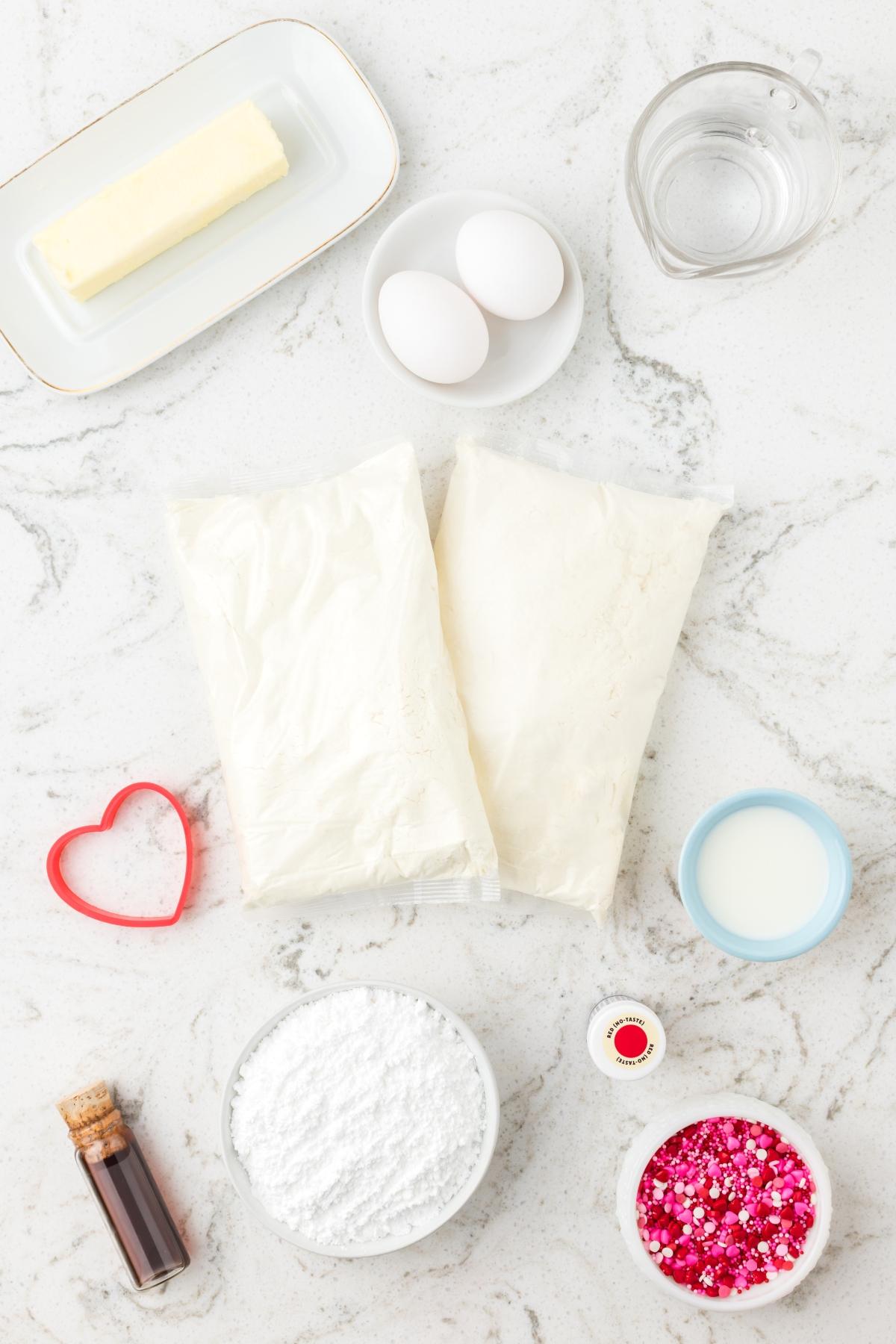 Butter, eggs, flour, sugar, milk, water, vanilla, sprinkles, and a heart-shaped cookie cutter on a countertop.