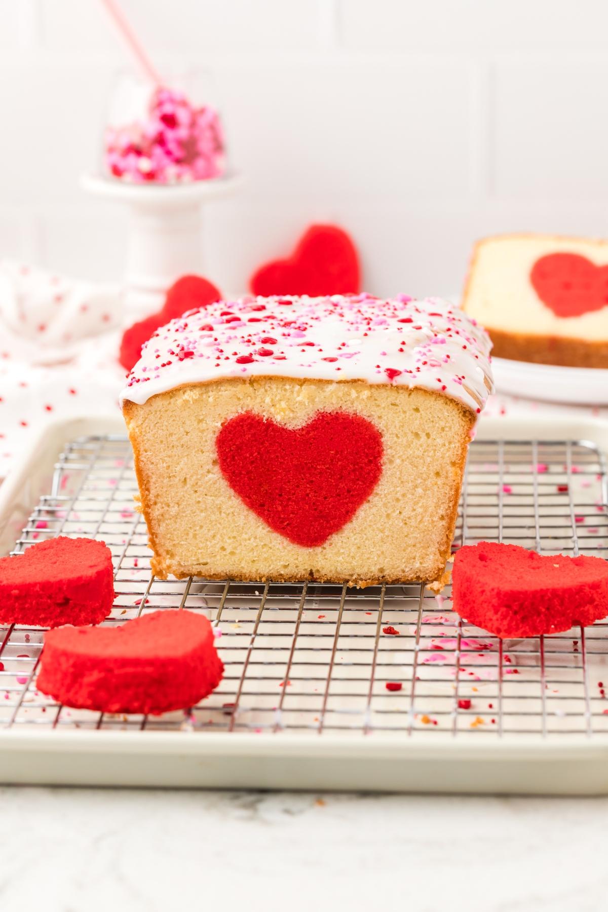 A loaf cake with a red heart shape inside, topped with white icing and pink sprinkles, sits on a cooling rack.