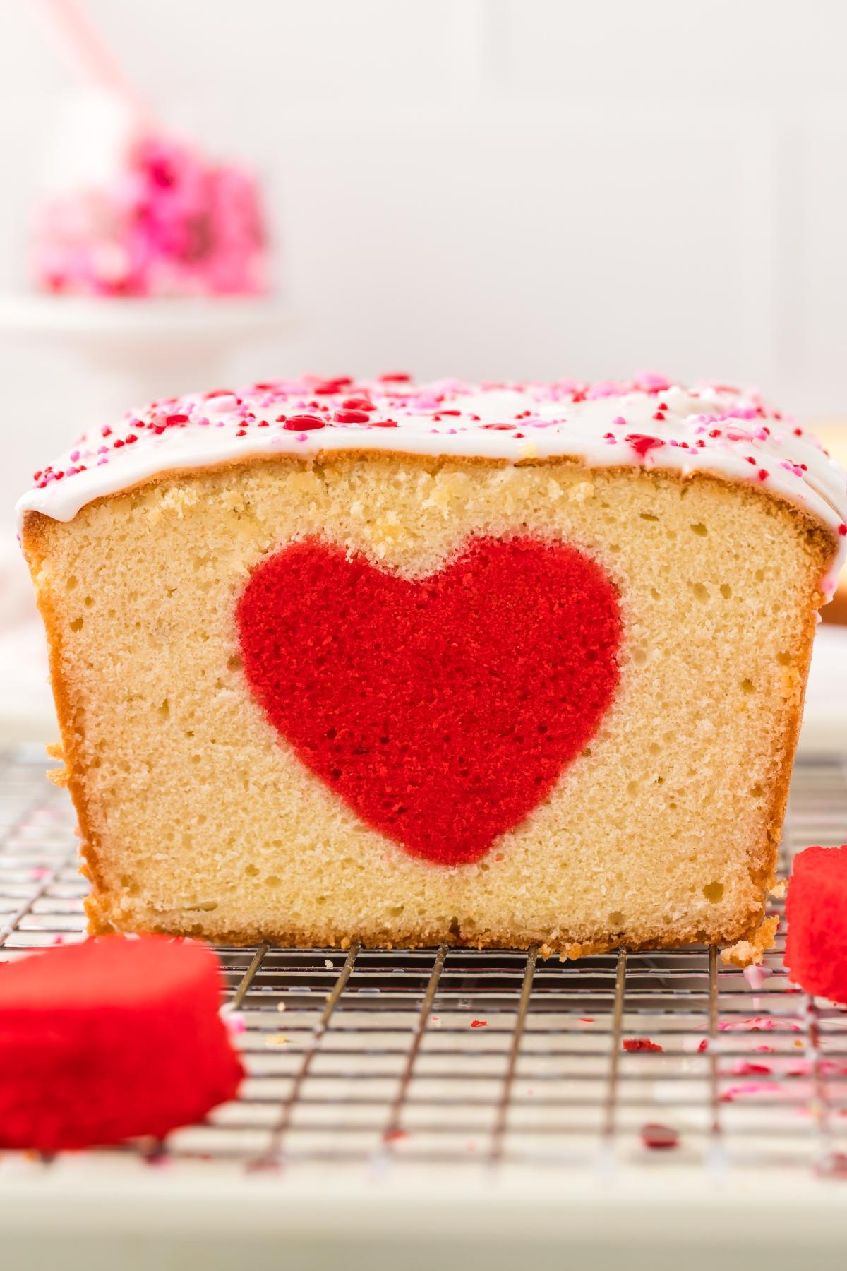 A loaf cake with a red heart shape inside, topped with white icing and pink sprinkles.