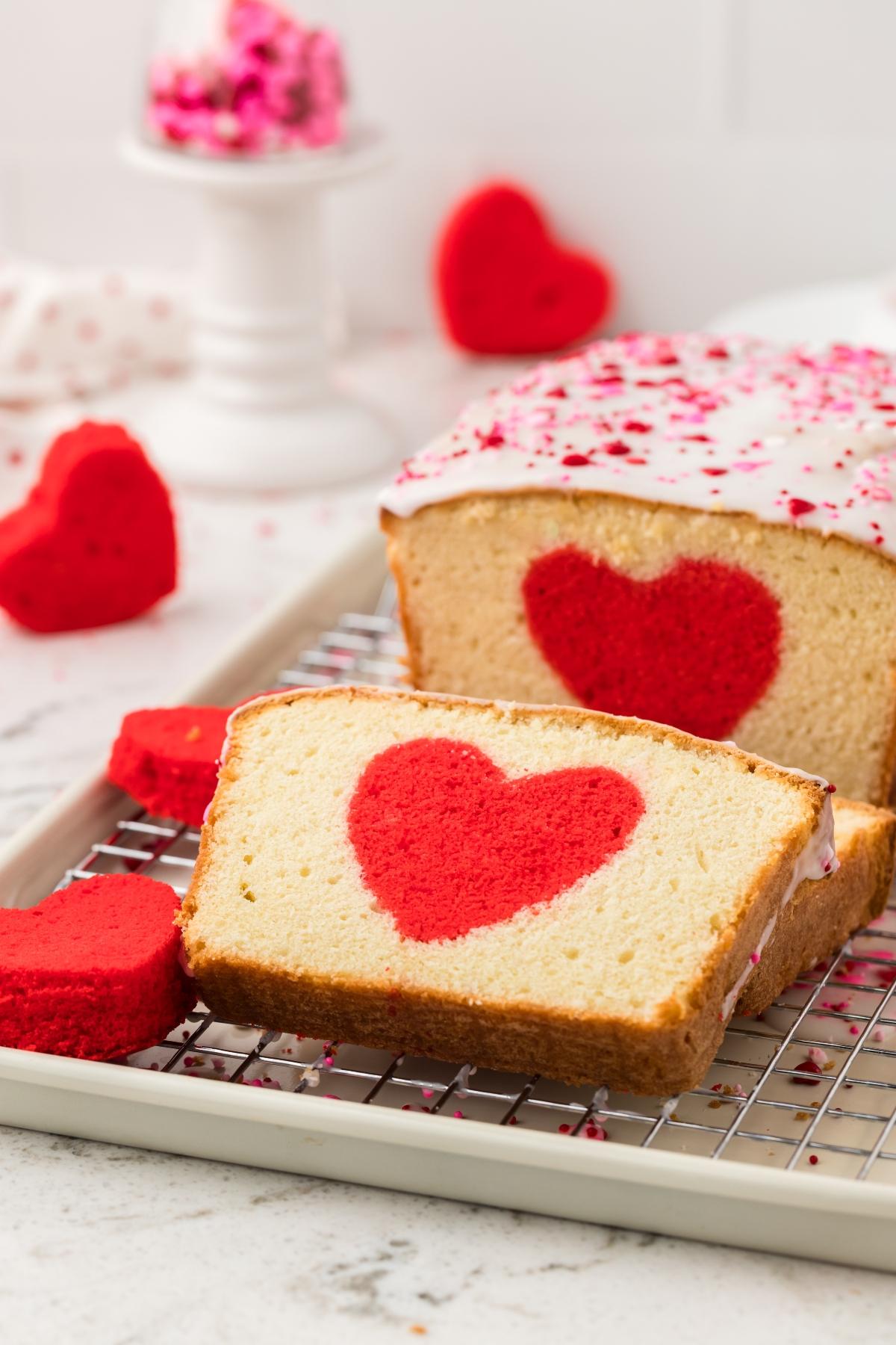 A loaf cake with a red heart shape inside, topped with white icing and pink sprinkles, on a cooling rack.