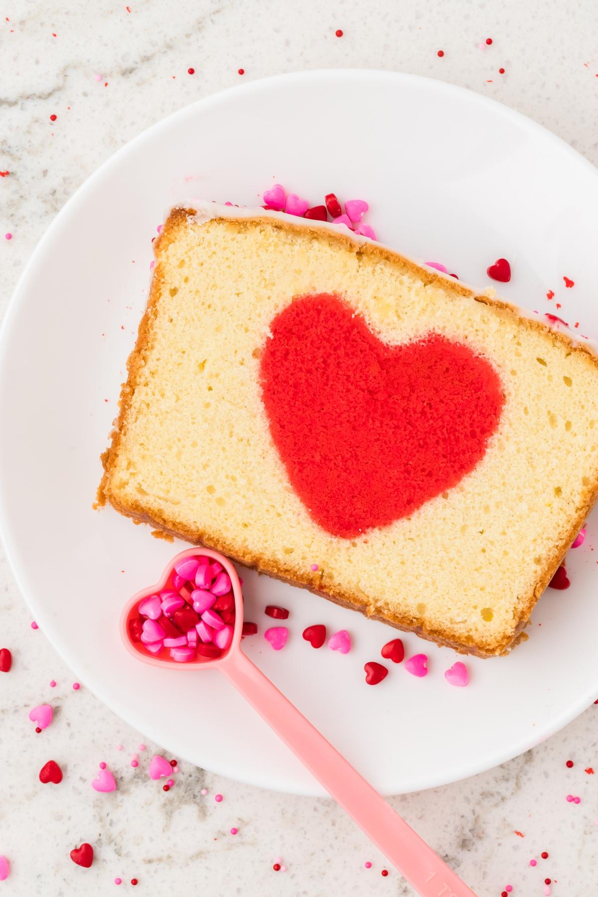 A slice of hidden heart cake with a red heart center, on a white plate with heart-shaped sprinkles and a pink spoon.