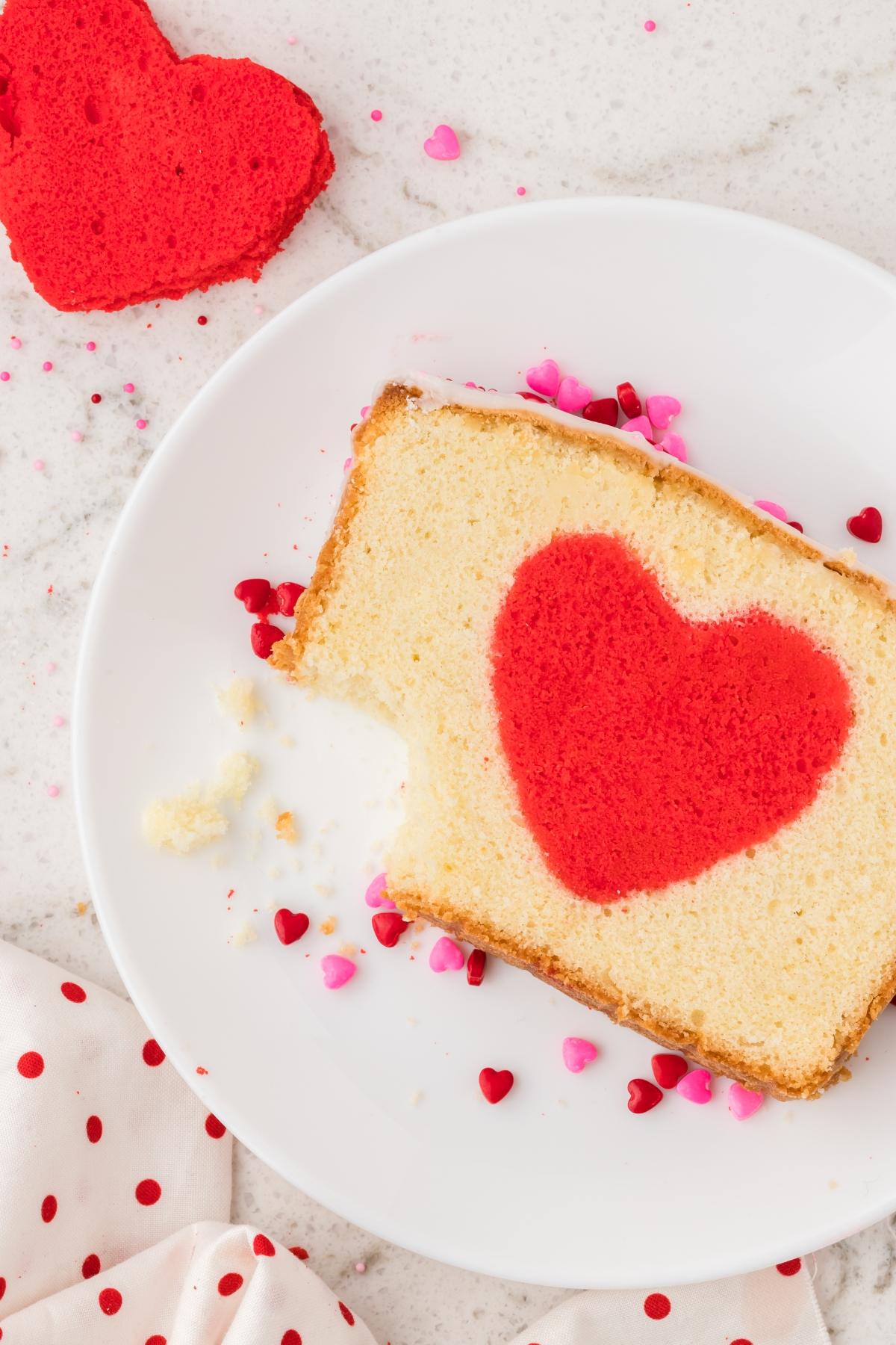 A slice of hidden heart cake with a red heart shape in the center, surrounded by small heart sprinkles on a plate.