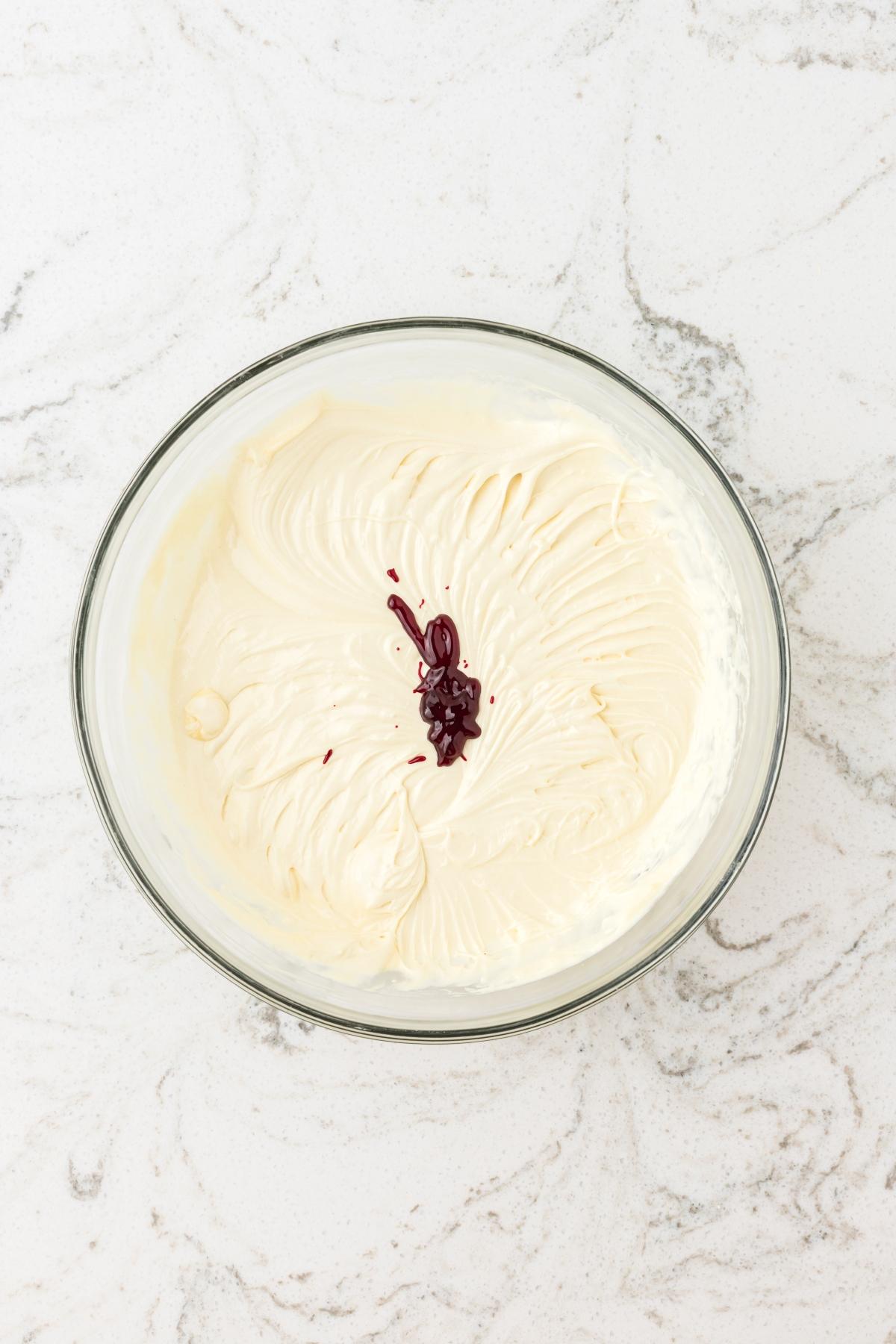 A glass bowl of whipped cream with a small amount of red liquid added on top, on a marble surface.