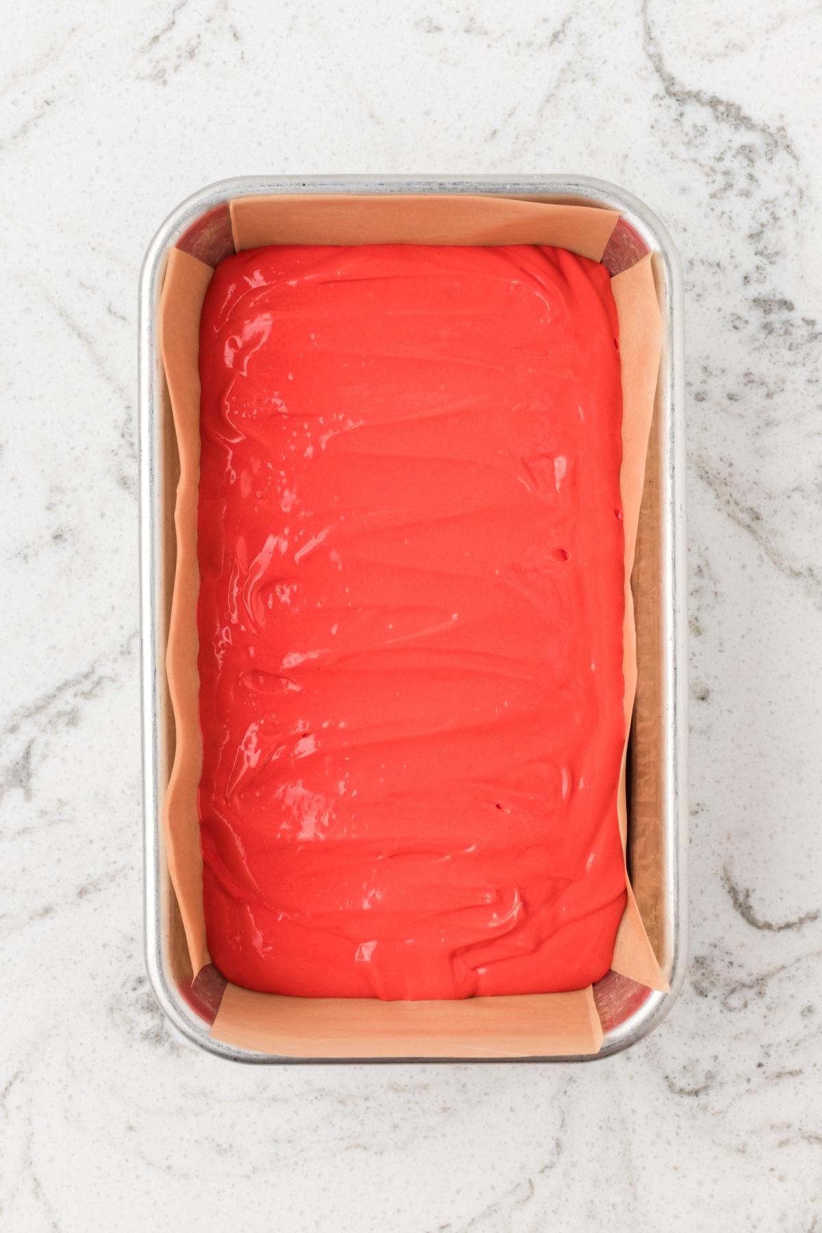 A loaf pan lined with parchment paper, filled with smooth, bright red batter on a marble surface.