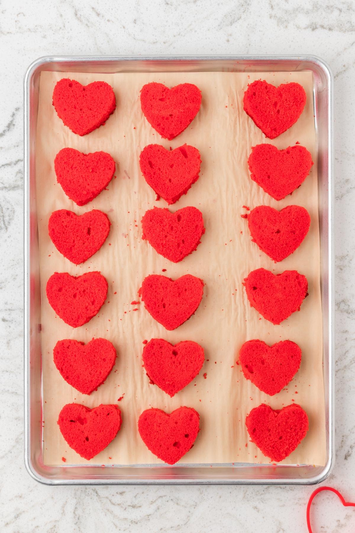 Red heart-shaped cake pieces arranged in rows on a parchment-lined baking sheet.