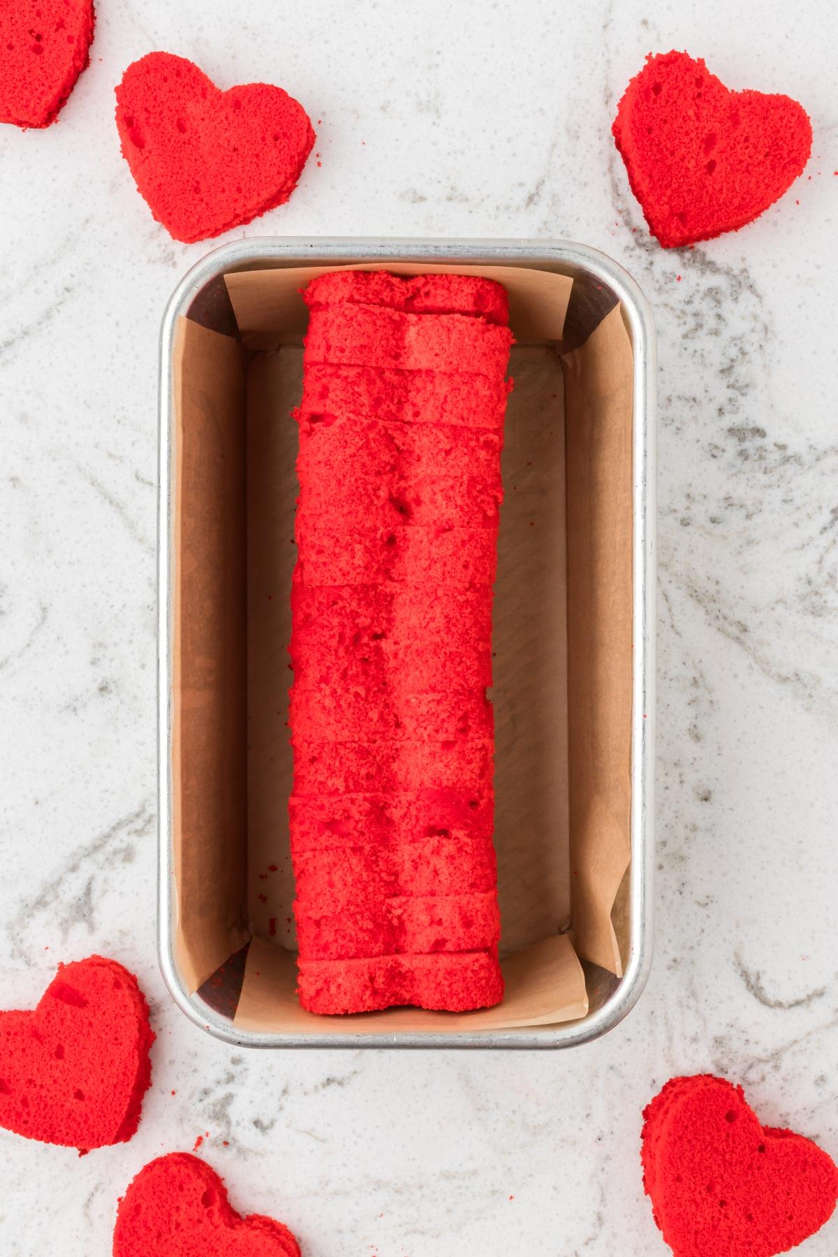 Bright red heart-shaped cake slices arranged in a loaf pan, with more heart cakes scattered around.