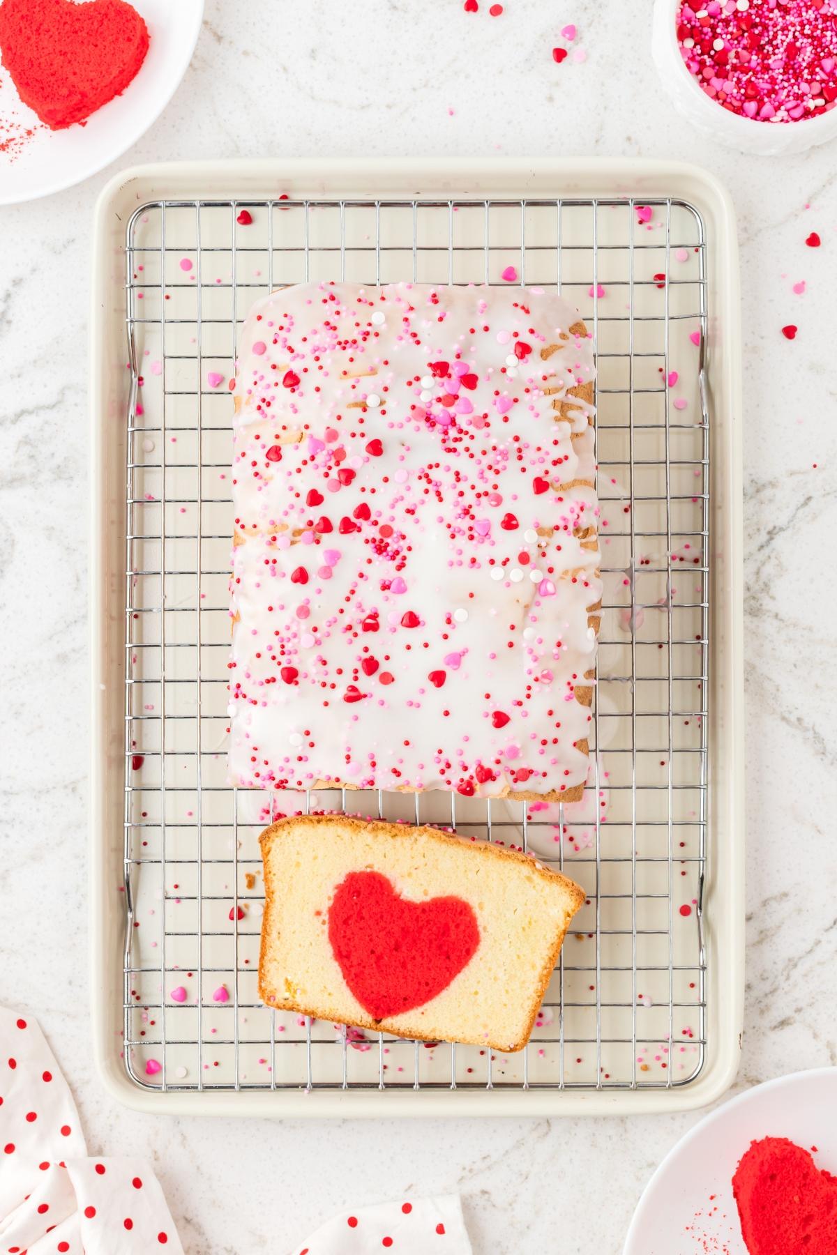 A loaf cake with white icing and heart sprinkles, one slice shows a red heart shape inside.