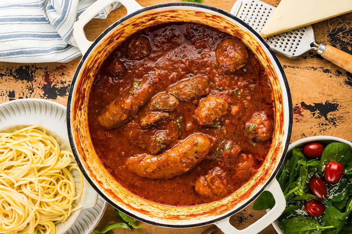 Large pot of Italian sausage in tomato sauce, just like a classic Sunday Sauce, with spaghetti, spinach, and Parmesan on the side.