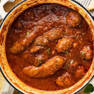 A pot of sausages simmering in rich Sunday Sauce beside a plate of spaghetti and a salad with cherry tomatoes.