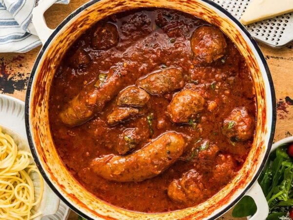 A pot of sausages simmering in rich Sunday Sauce beside a plate of spaghetti and a salad with cherry tomatoes.