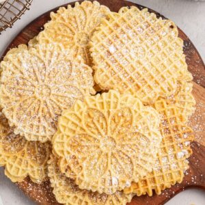Round, patterned Italian Pizzelle cookies dusted with powdered sugar on a wooden board, with some cookies set off to the side.