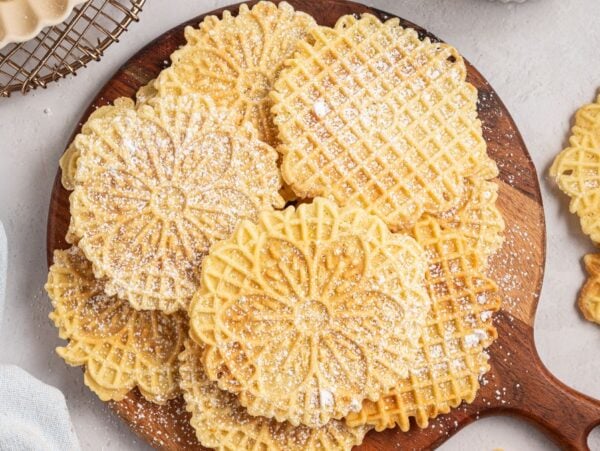 Round, patterned Italian Pizzelle cookies dusted with powdered sugar on a wooden board, with some cookies set off to the side.