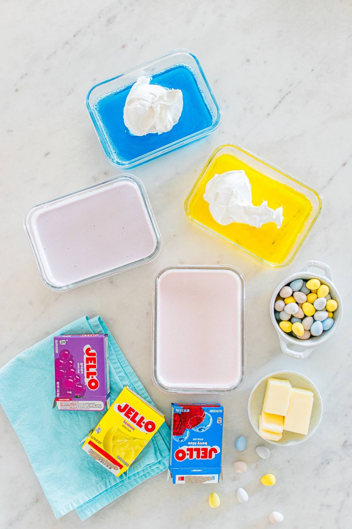 Three dishes of pastel Jello, Cool Whip, candy eggs, butter, and Jello boxes on a marble surface.