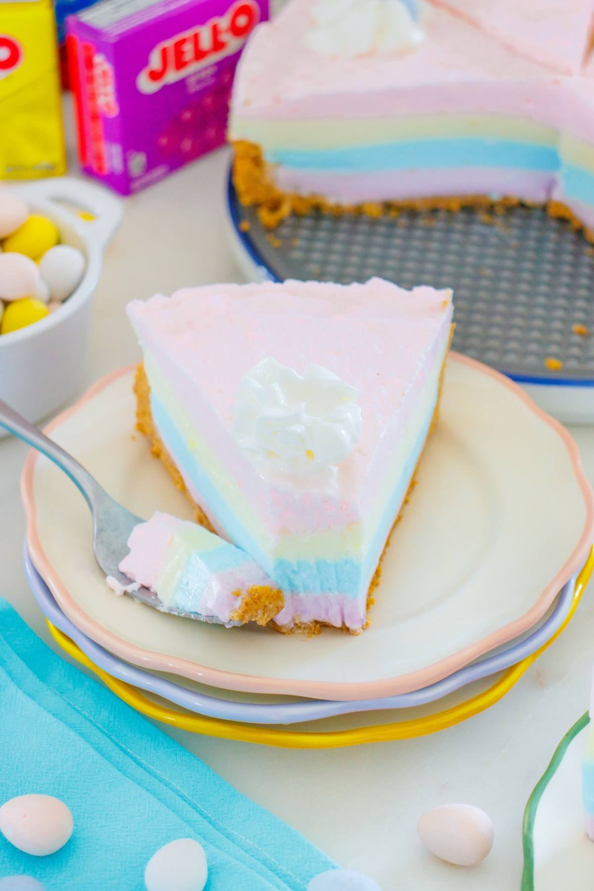 A layered jello pie cheesecake slice with whipped cream on top, on a plate next to a fork and colorful candies.