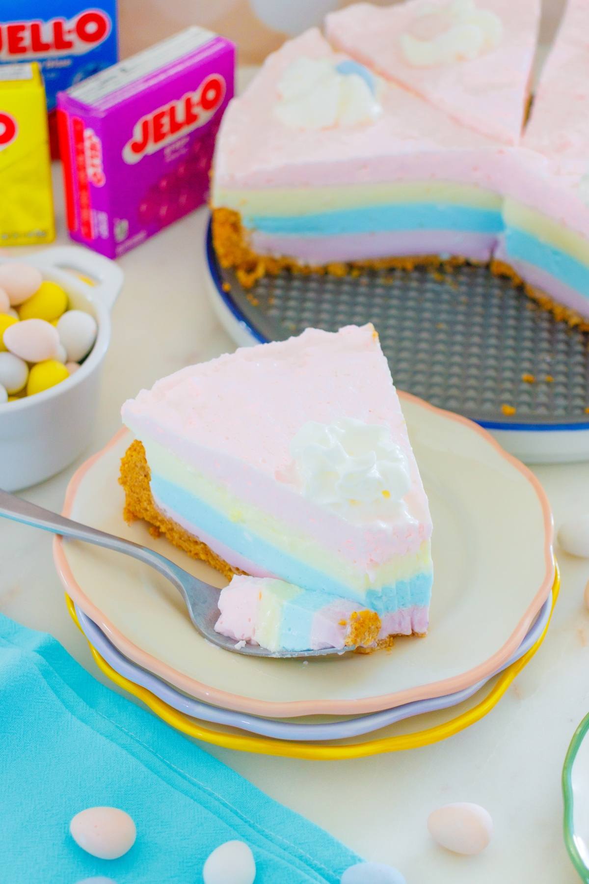 A jello pie cheesecake slice with whipped cream on a plate, next to a whole cheesecake and Jello boxes.