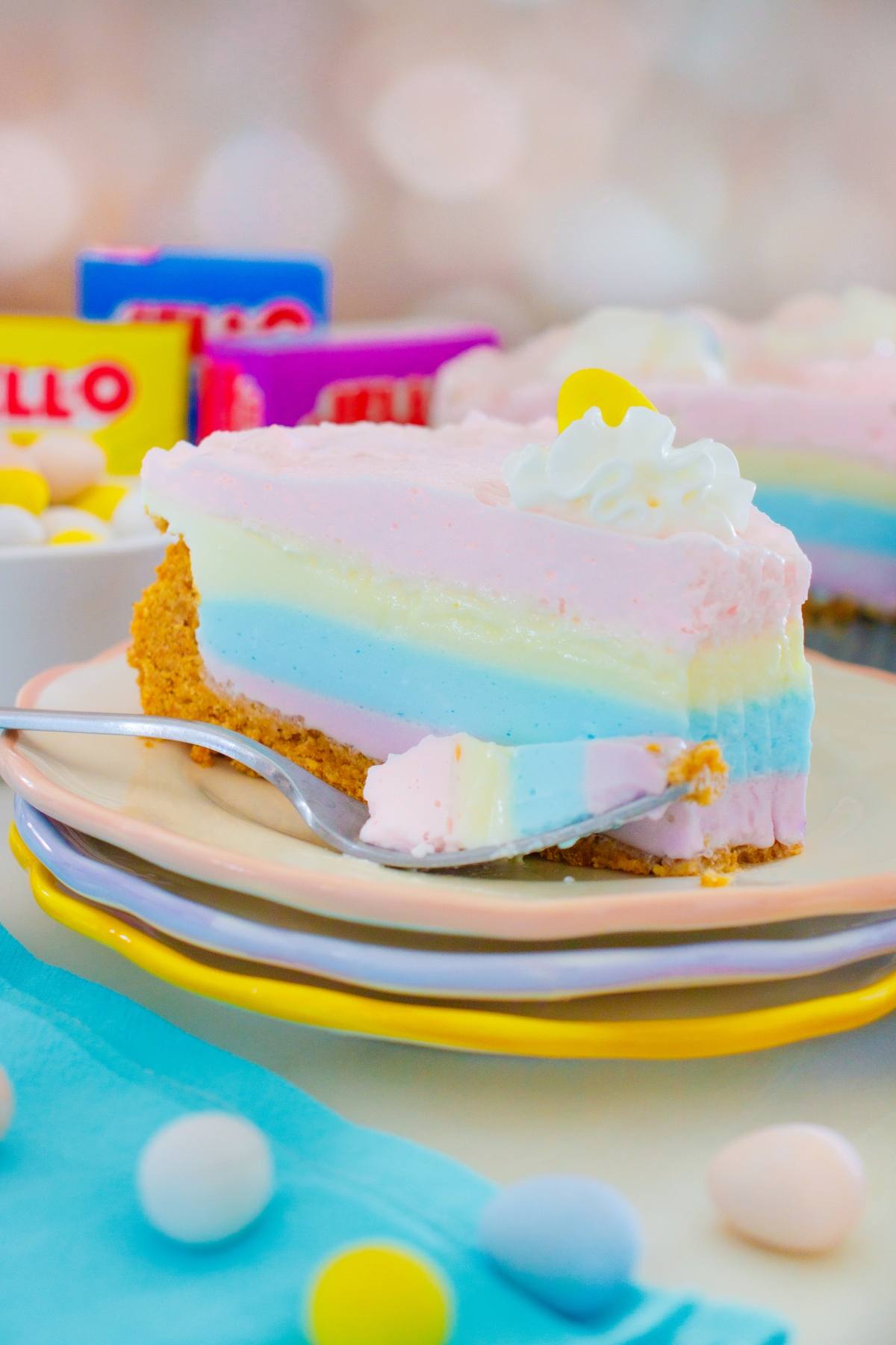 A slice of layered jello pie  cheesecake on a plate with a fork, surrounded by candy and Jello boxes.