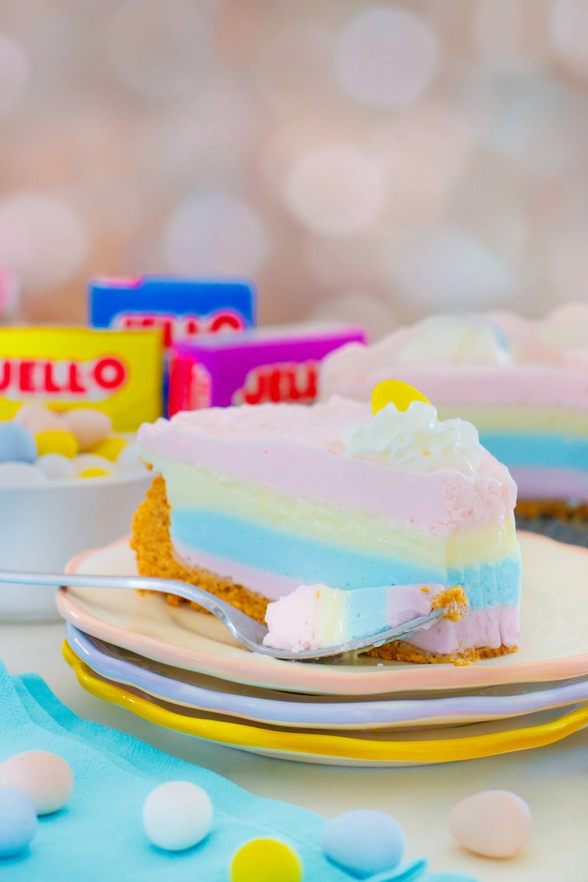 A layered jello pie cheesecake slice on a plate with candy eggs and Jello boxes in the background.