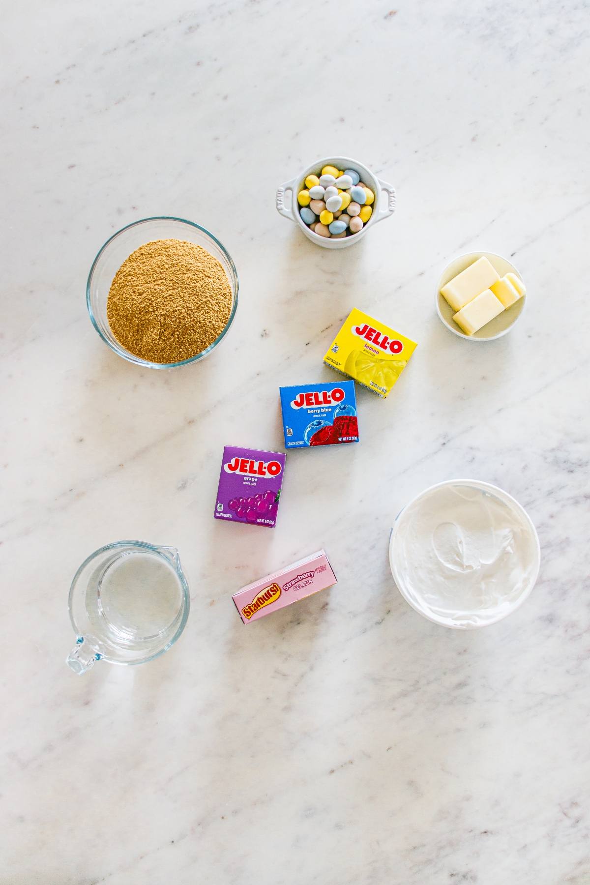 Baking ingredients on a marble counter: graham crumbs, butter, candy, water, whipped topping, and Jello boxes.