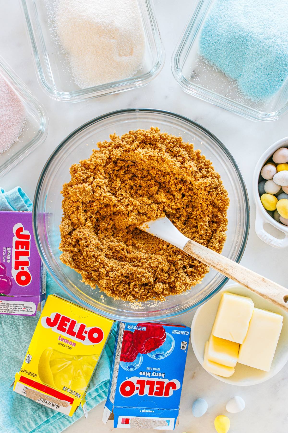 A bowl of graham cracker crust mix with a spatula, Jello boxes, butter, sugar, and mini eggs on the counter.
