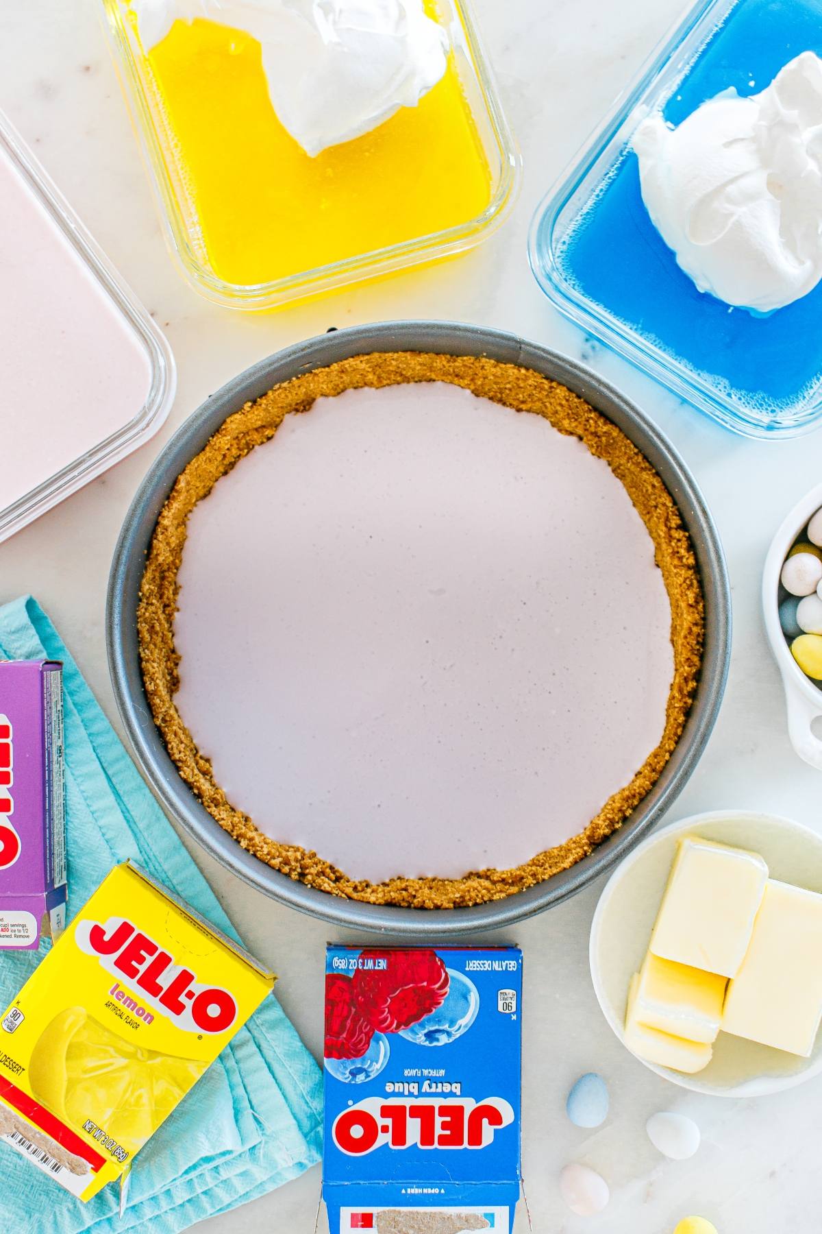 Round pie with graham crust surrounded by Jello, whipped topping, butter, and colorful candies on a table.