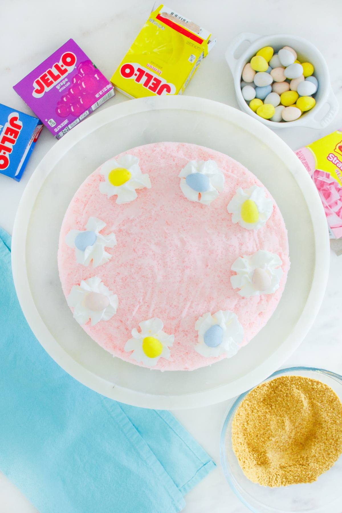A pink Jello dessert on a white plate, topped with whipped cream and candy eggs, surrounded by boxes of Jello.