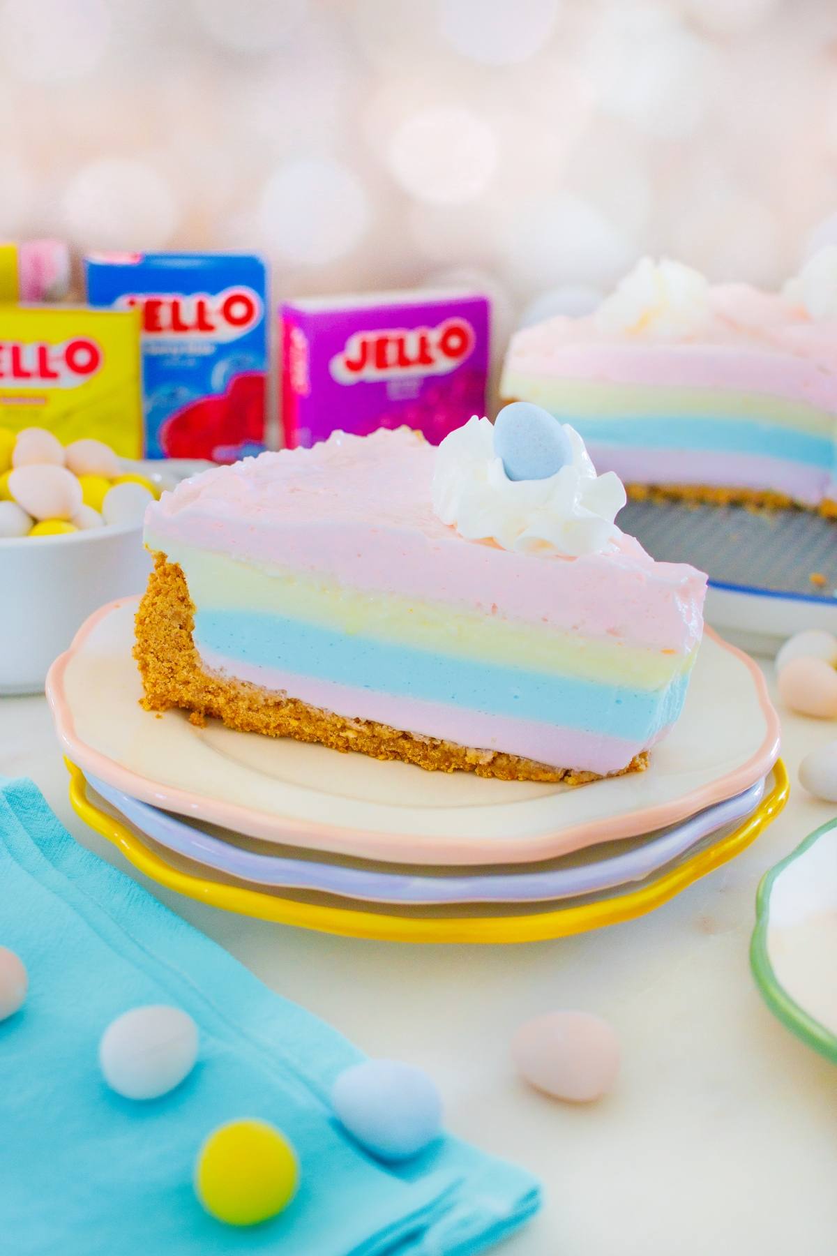 A jello pie rainbow cheesecake slice with whipped cream and a candy egg, surrounded by colorful candies and Jell-O boxes.