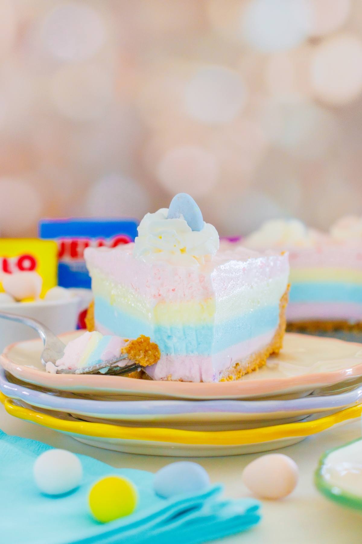 A layered jello pie cheesecake slice with whipped cream and a candy egg on top, on a pastel plate.