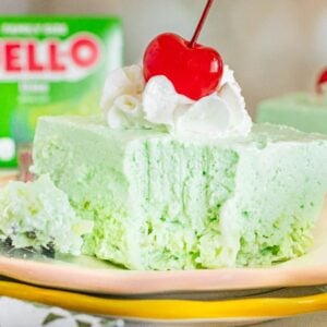 A slice of light green Lime Jello Salad topped with whipped cream and a cherry sits on a plate, with a Jell-O box in the background.