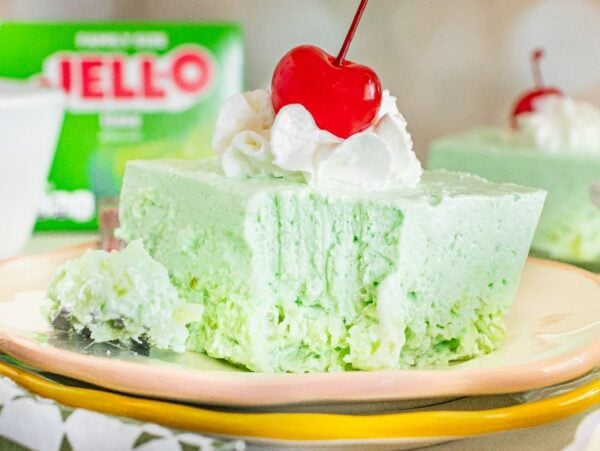A slice of light green Lime Jello Salad topped with whipped cream and a cherry sits on a plate, with a Jell-O box in the background.