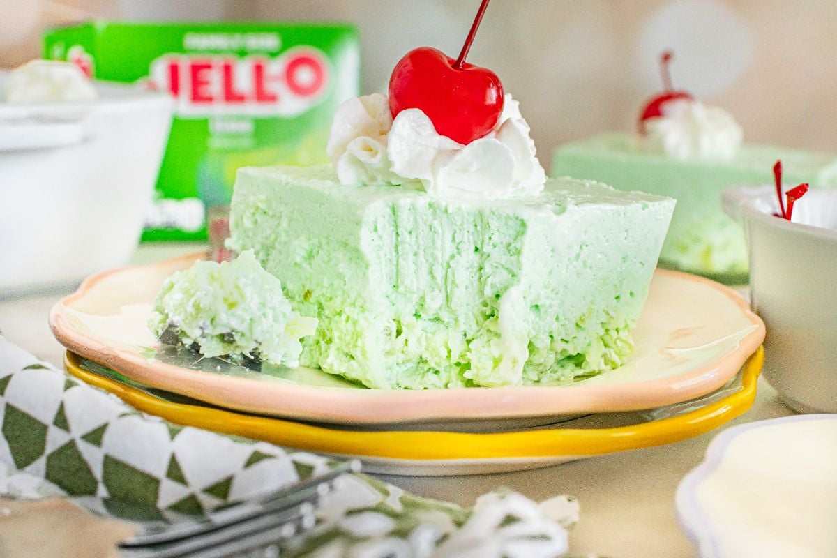 A slice of light green Lime Jello Salad topped with whipped cream and a cherry sits on a plate.