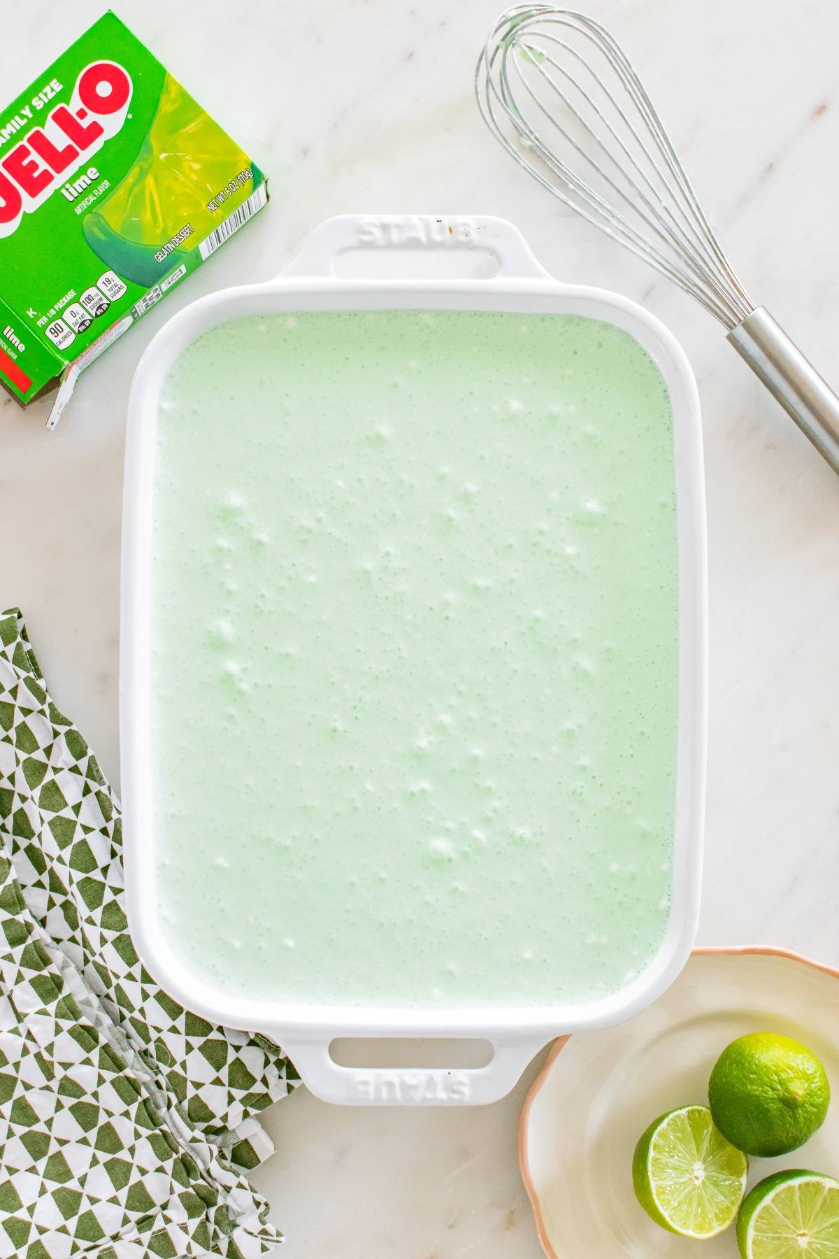A white dish filled with light green Jello mixture next to a whisk, limes, and a box of lime Jello.