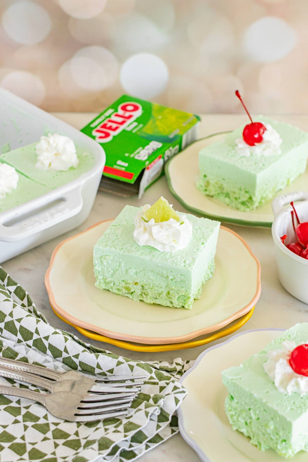 Light green Jello salad dessert squares topped with whipped cream and cherries on plates, with Jello box in background.