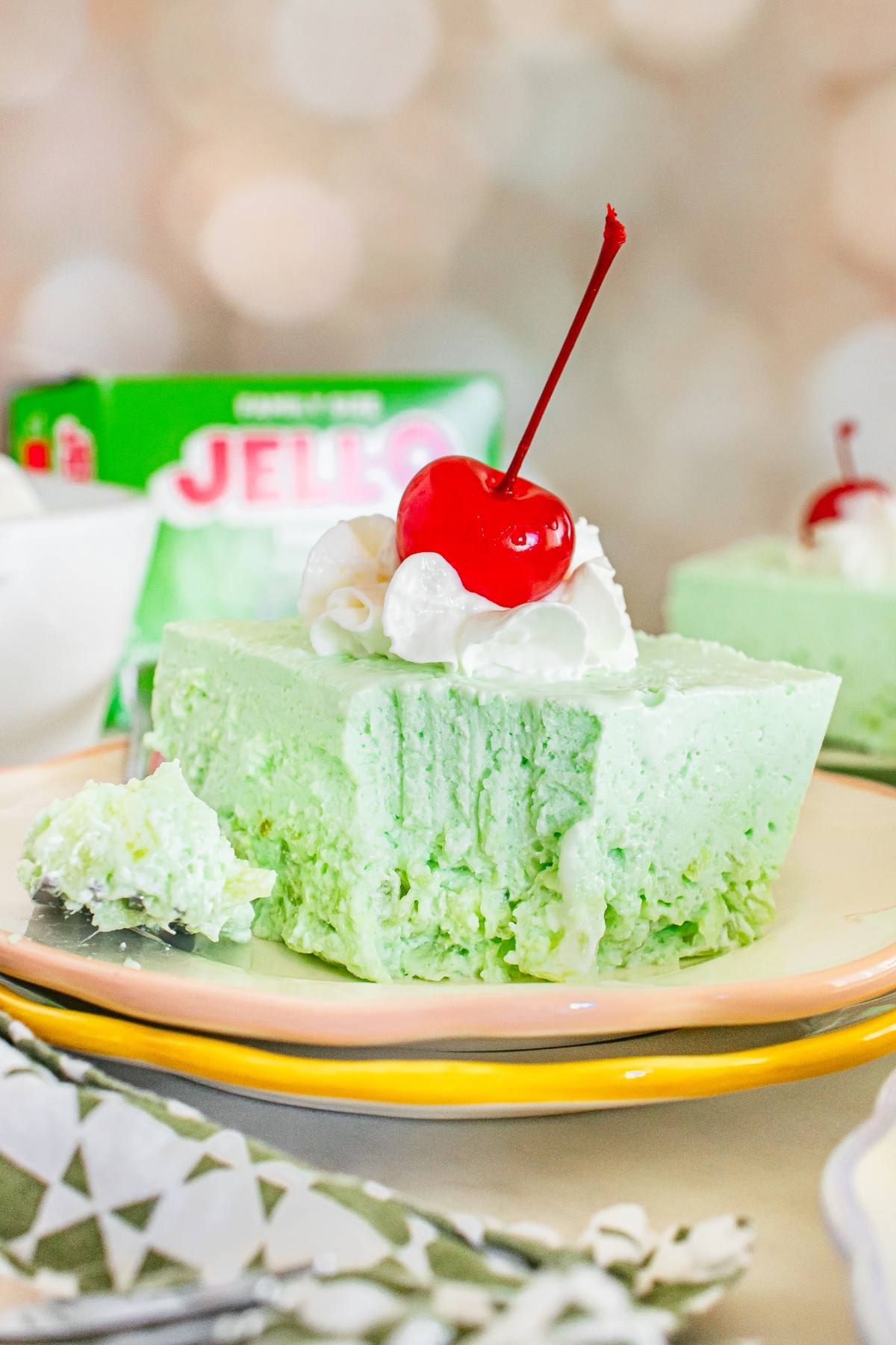 A slice of lime Jello salad cheesecake topped with whipped cream and a cherry, with a bite taken out.