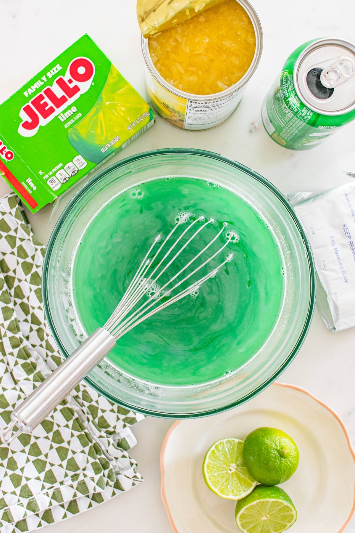 A bowl of green Jell-O mix with a whisk, surrounded by lime, soda, cream cheese, and pineapple.