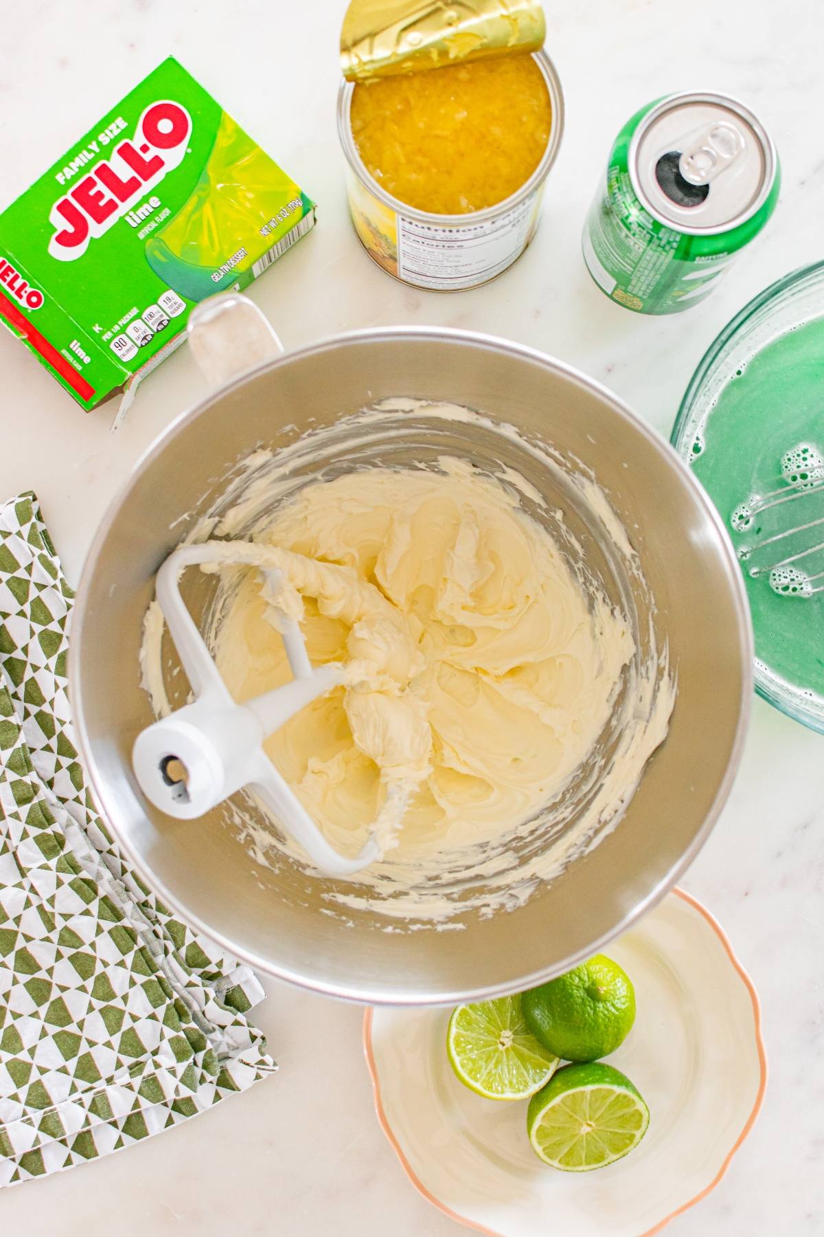 Mixing bowl with creamy batter, lime Jello box, canned pineapple, lime, and green soda on a white surface.
